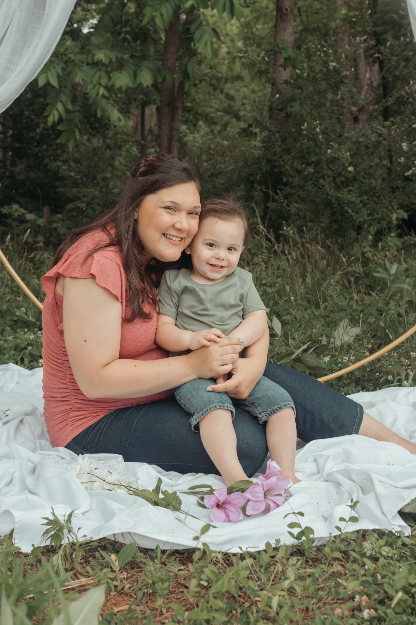 Parent and child sitting on white blanket outdoors during casual photo session in wooded area.
