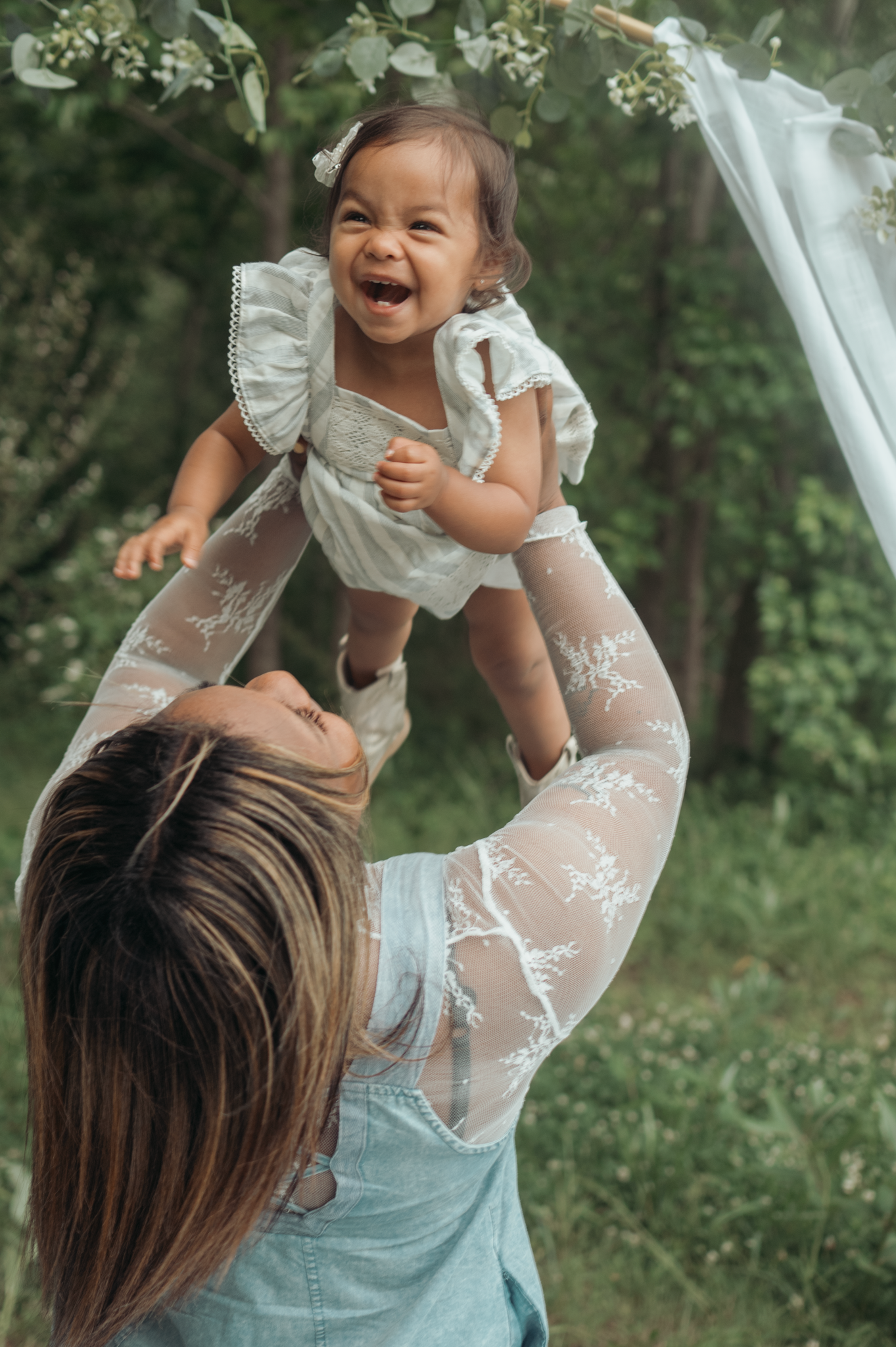 A joyful moment captured outdoors as a baby wearing a white dress is lifted up high in a garden setting.