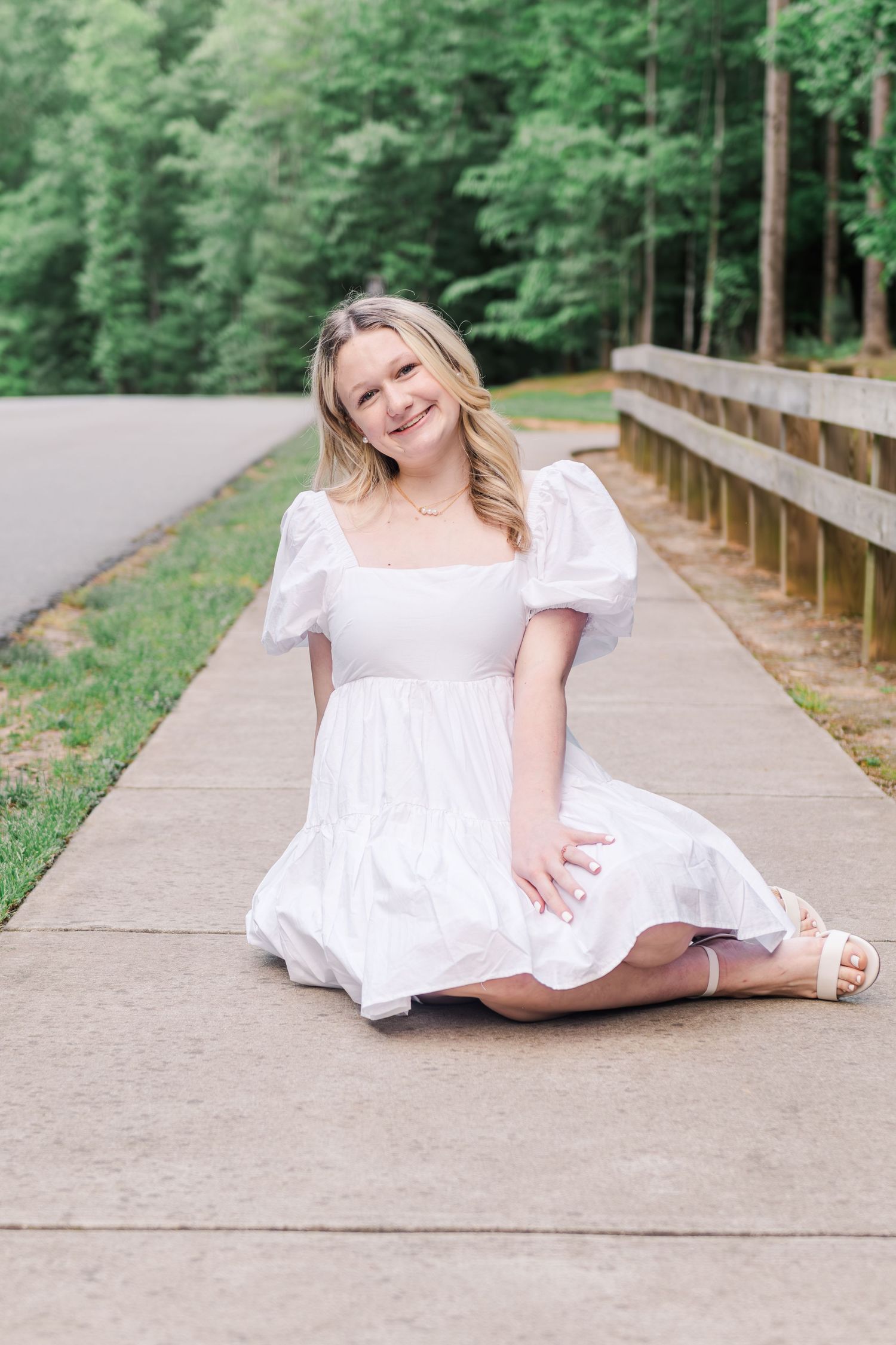 A person in a white summer dress sits on a wooden boardwalk path surrounded by lush green forest.