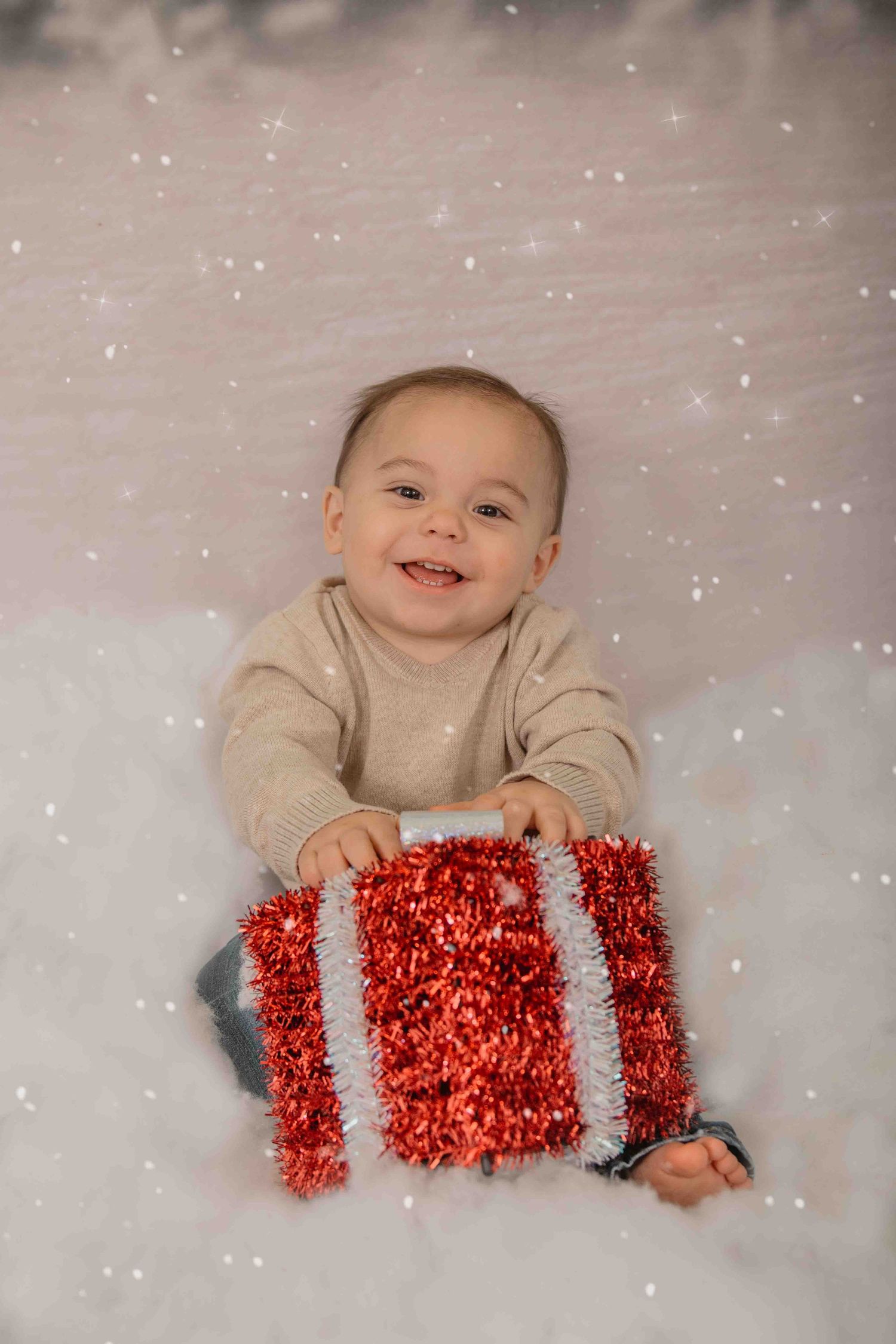 Baby in beige sweater lying on white backdrop with sparkly overlay holding red tinsel wrapped present box.