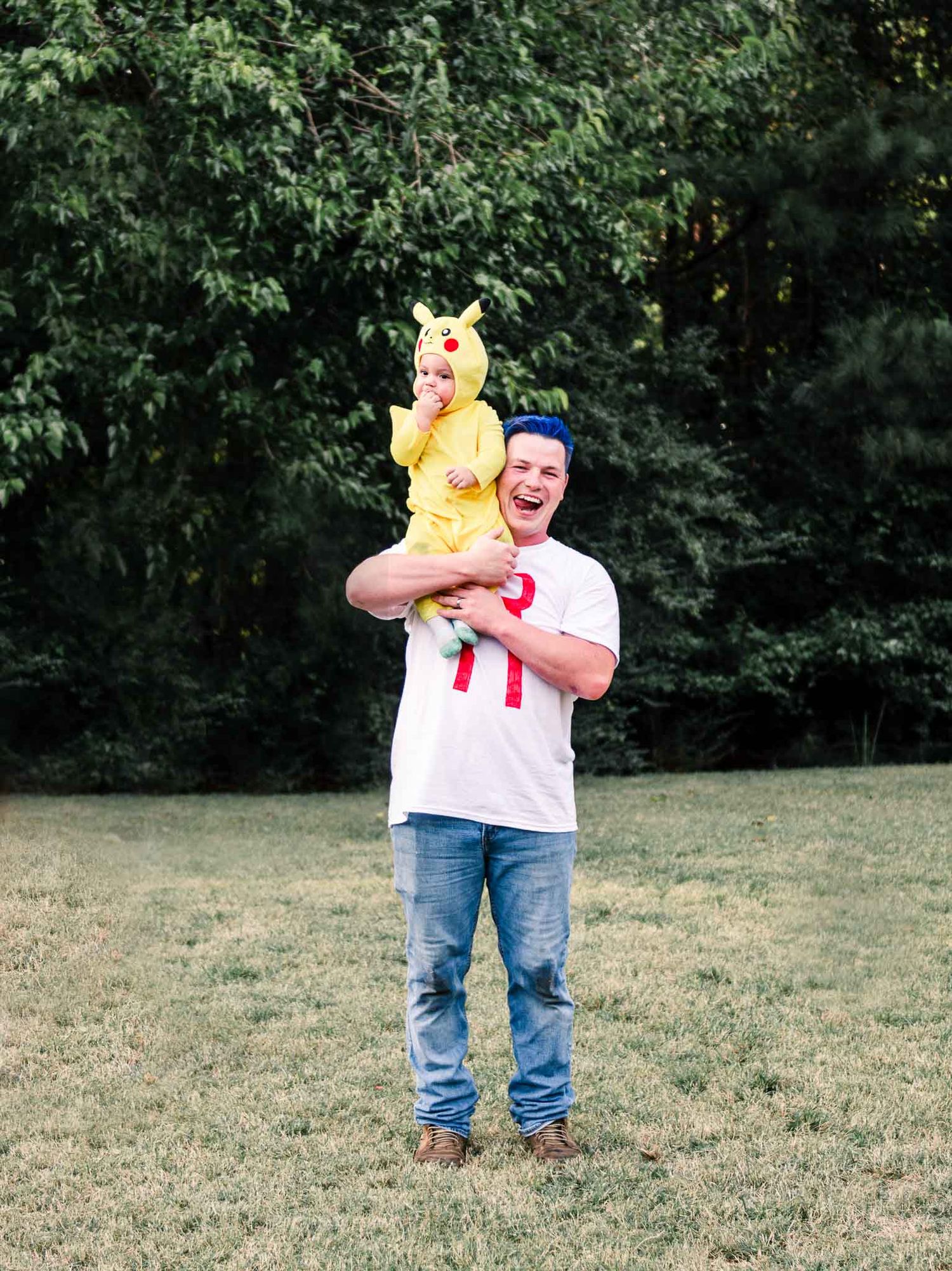 Someone wearing a white t-shirt and jeans holds a small dog dressed in a yellow Pikachu costume in an outdoor setting.