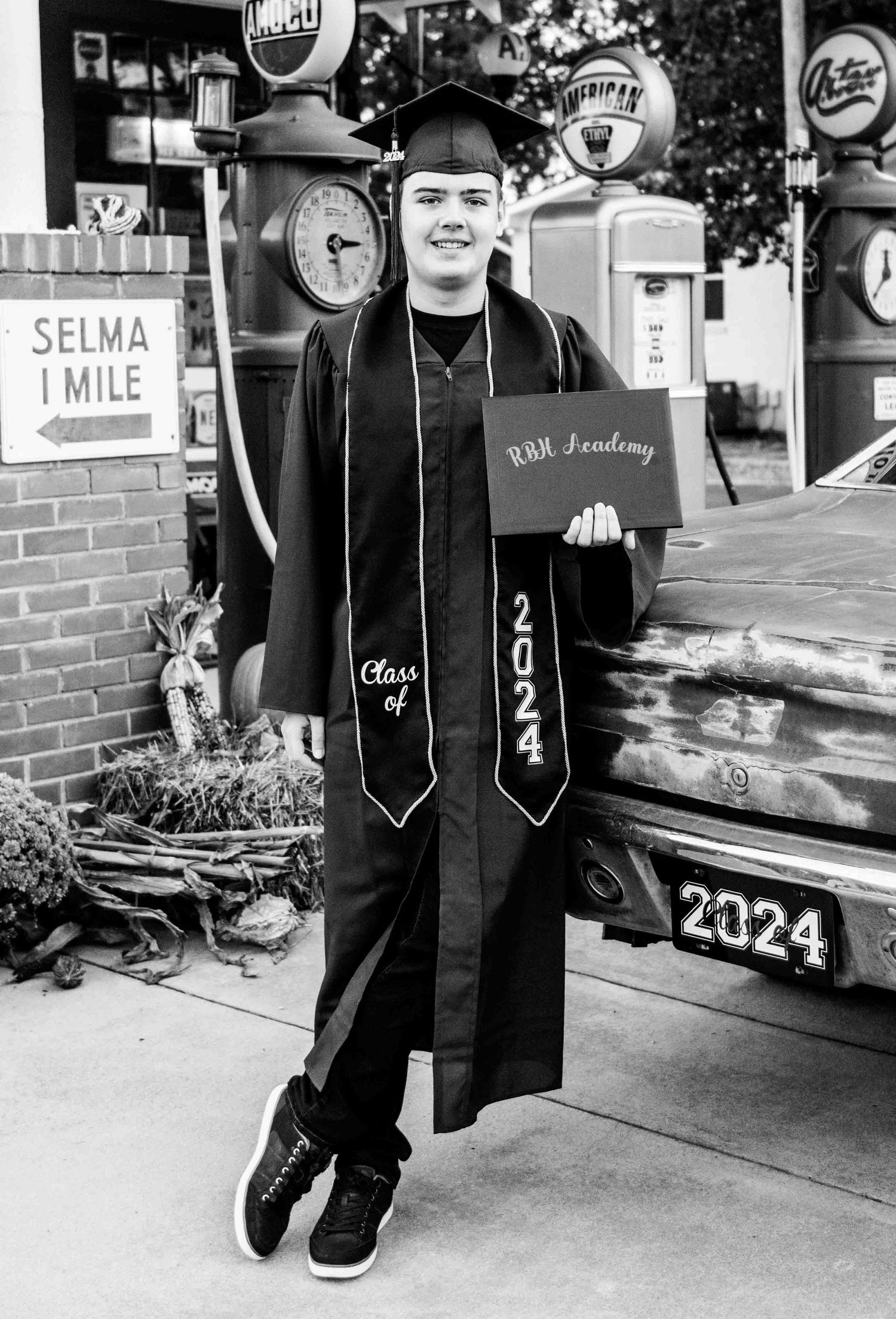 A black and white photo at a vintage gas station showing a graduate in cap and gown posing with class of 2024 regalia.