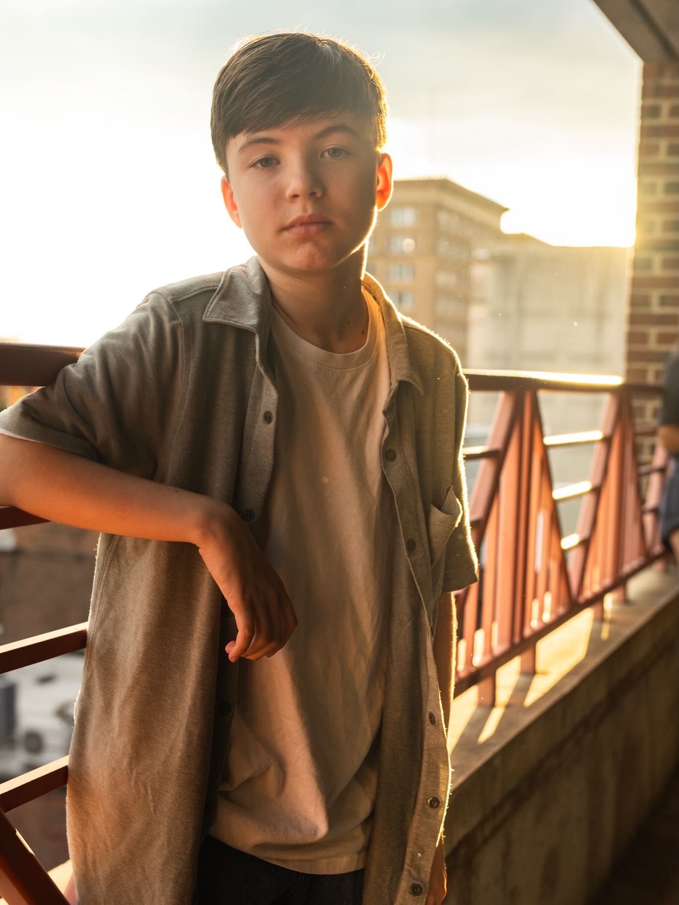 A young boy leans on a railing at golden hour, wearing a gray shirt over a white tee with a city backdrop.