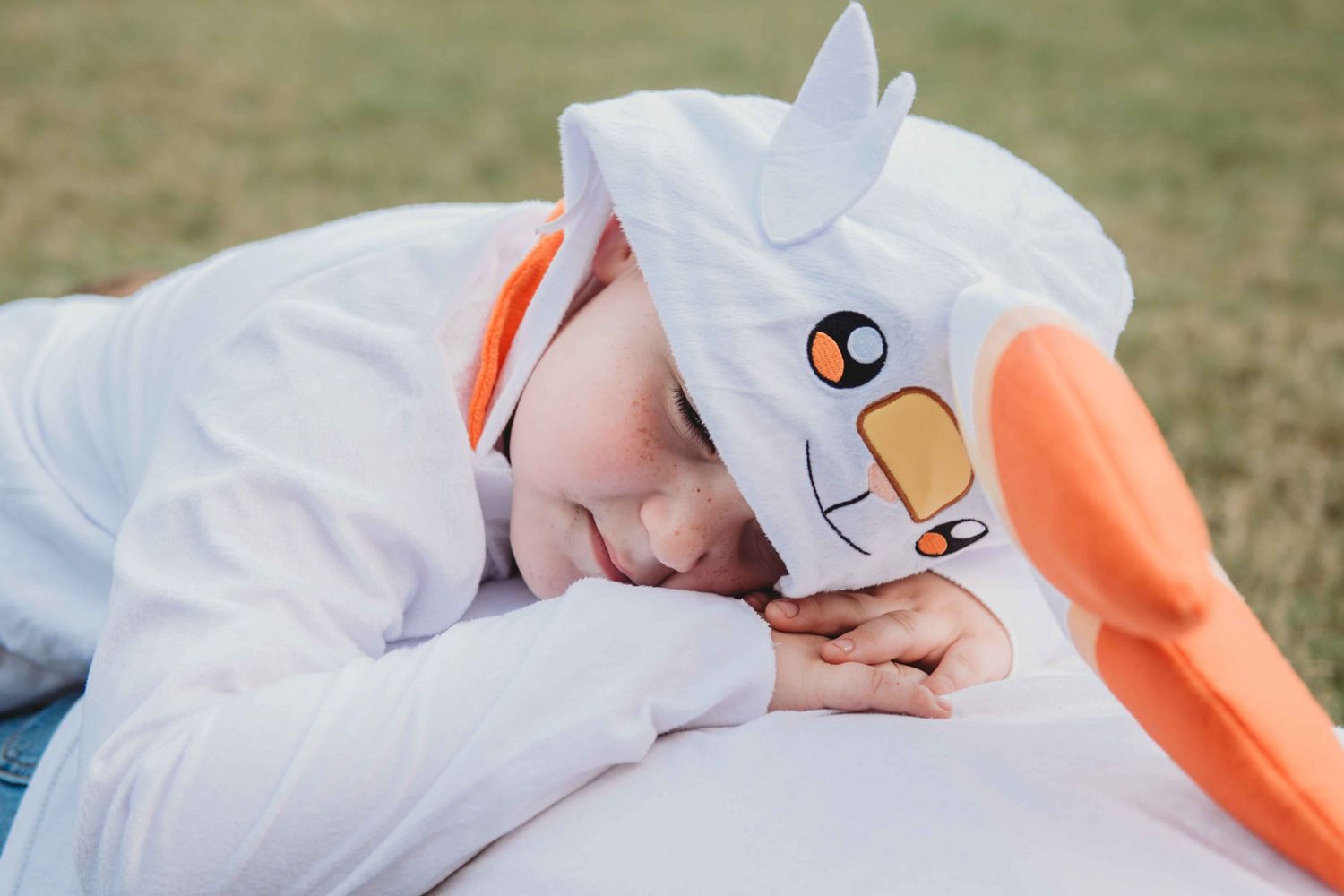 Someone dressed in a plush white swan costume with orange beak rests peacefully on green grass.