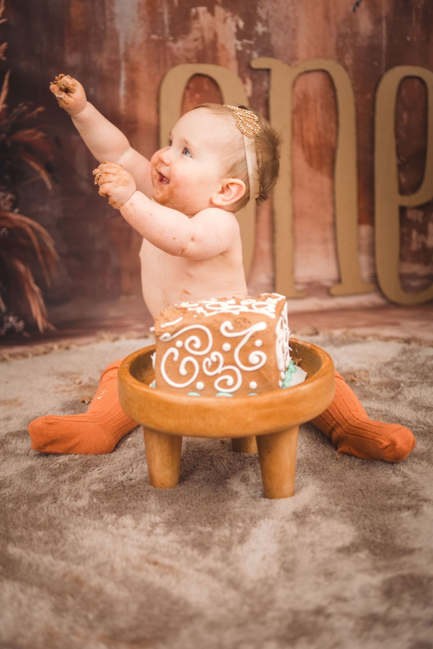 Baby sitting in decorative wooden stool wearing cake-inspired outfit during photoshoot with rustic background.