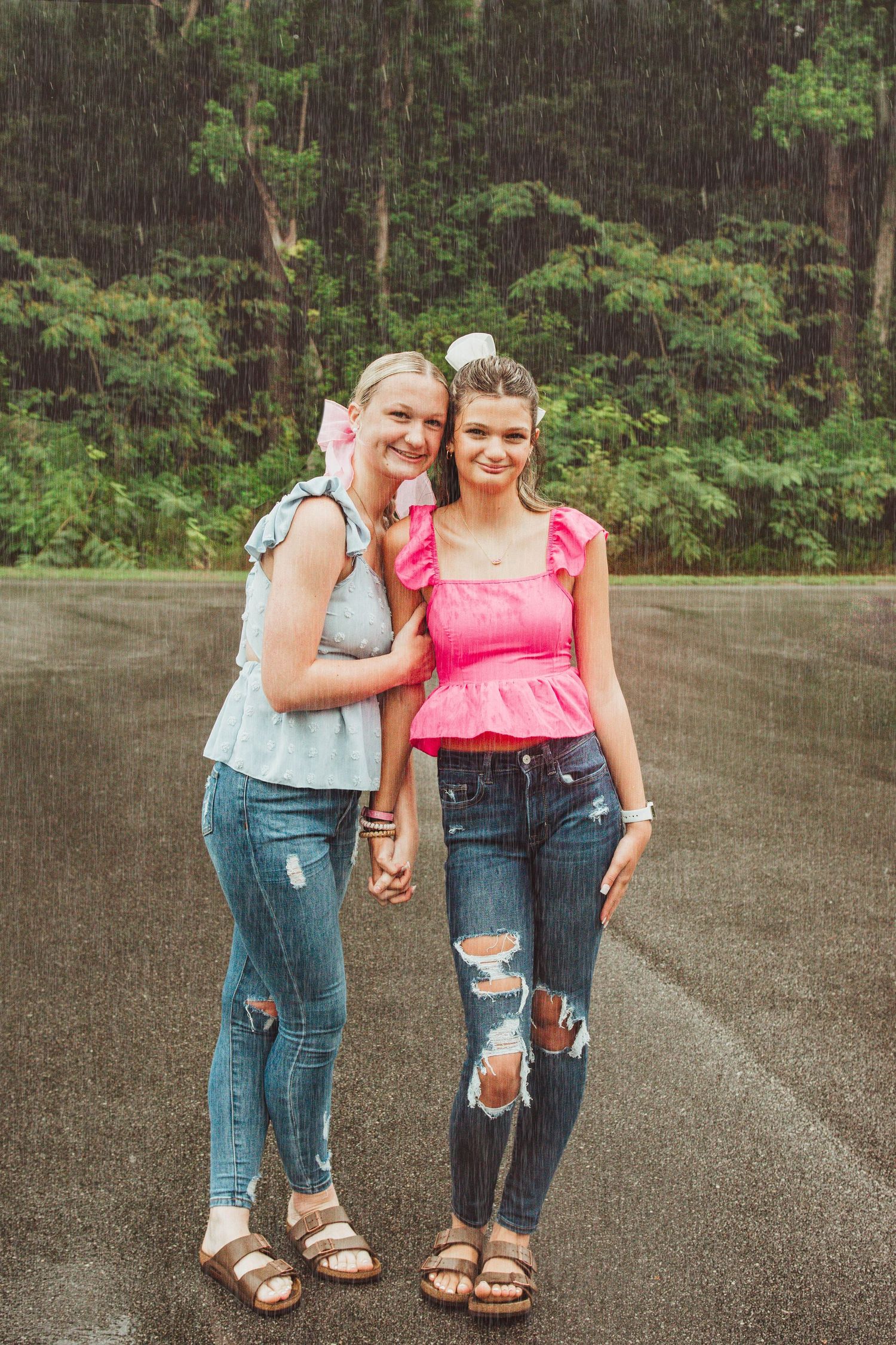 Two companions in casual summer attire stand together on a wet road with forest trees in the background.