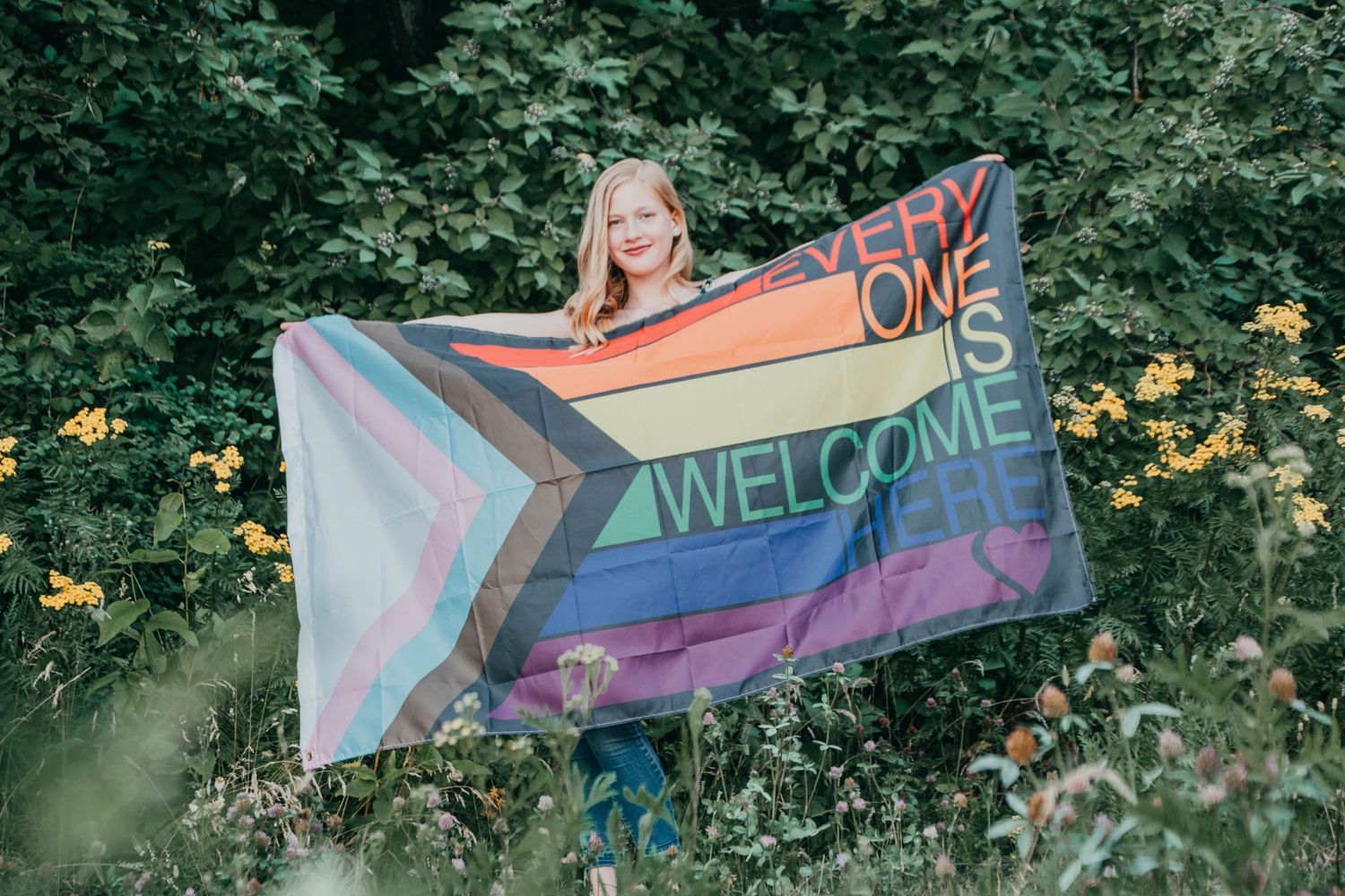 Someone stands in a garden holding a rainbow-colored Welcome flag featuring trans pride colors and stripes.