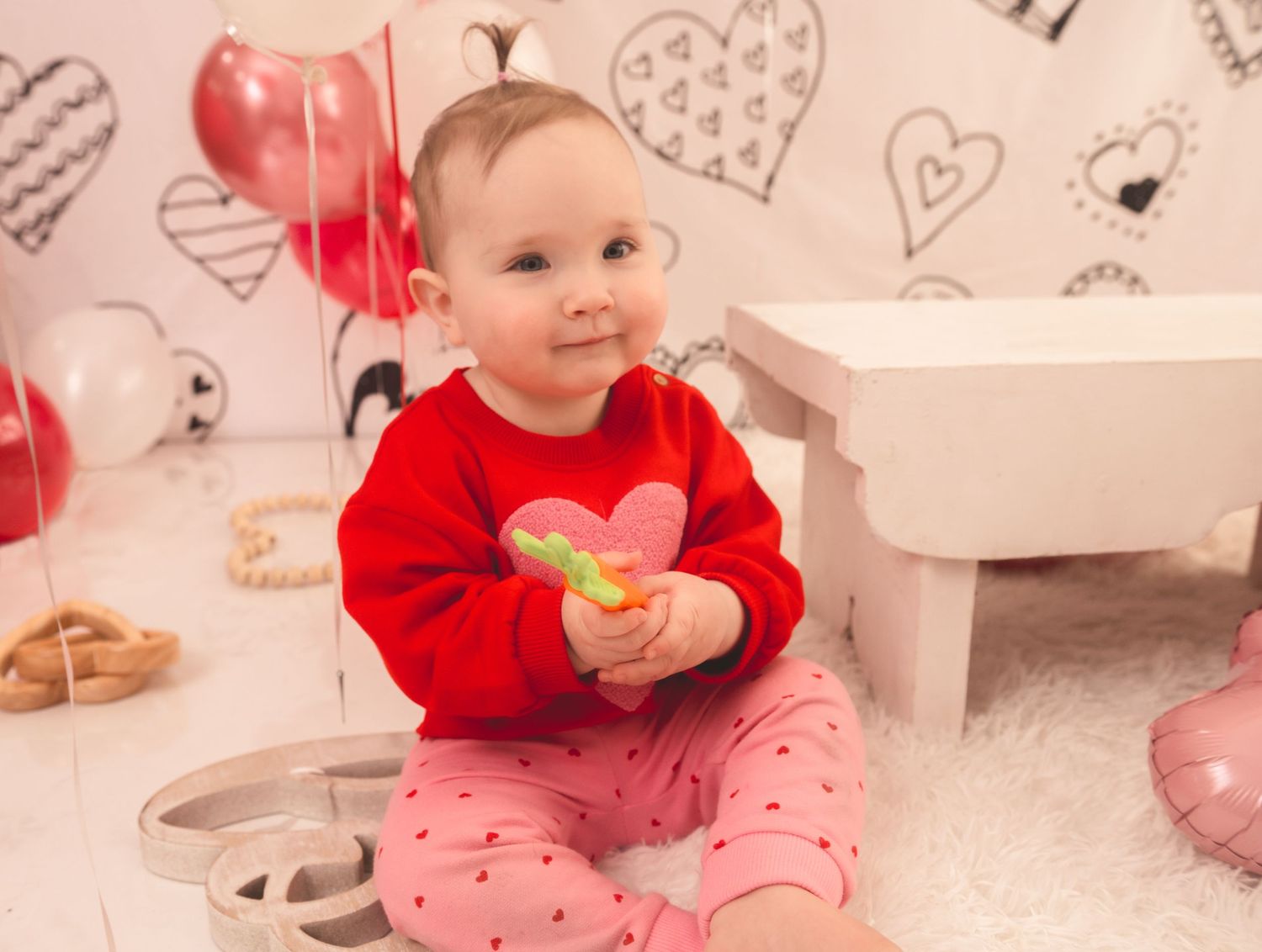 Small child wearing red sweater and pink heart pants sits on floor with Valentine's Day decorations around.