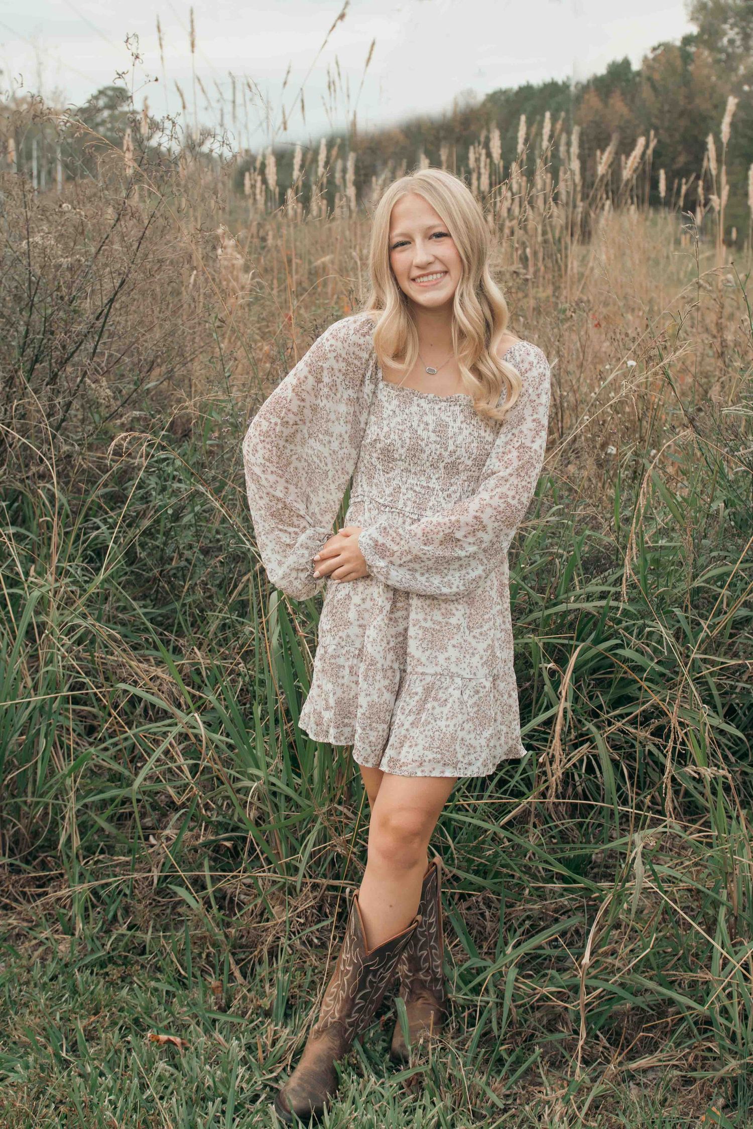 Woman in floral dress and boots poses in field of tall grass during golden hour.