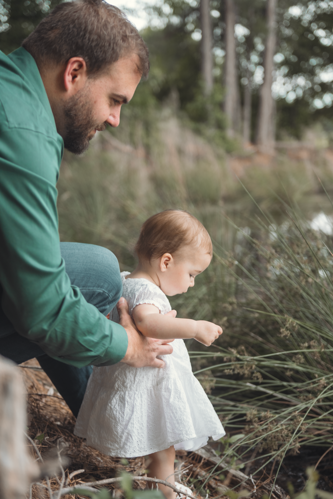 An adult in a green shirt carefully supports a small toddler in a white dress exploring tall grasses outdoors.