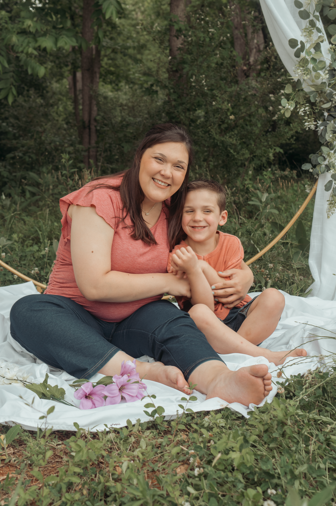 Outdoor portrait of two people sitting together on a white blanket with flowers and decorative arch in a forest setting.