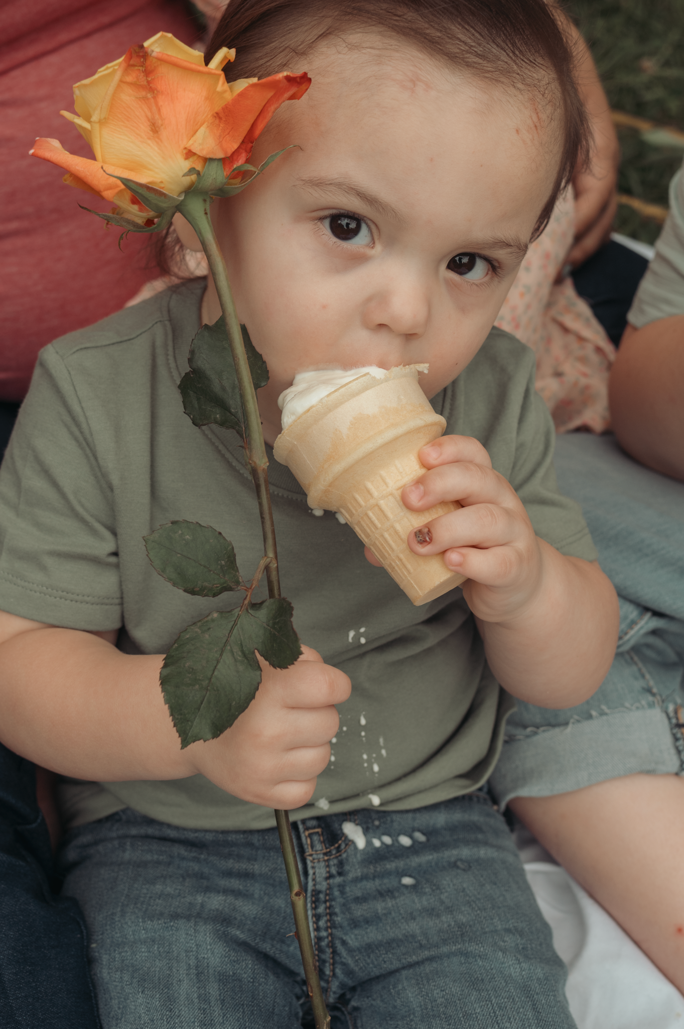 Child in green shirt holds an orange rose while enjoying vanilla ice cream cone on summer day.