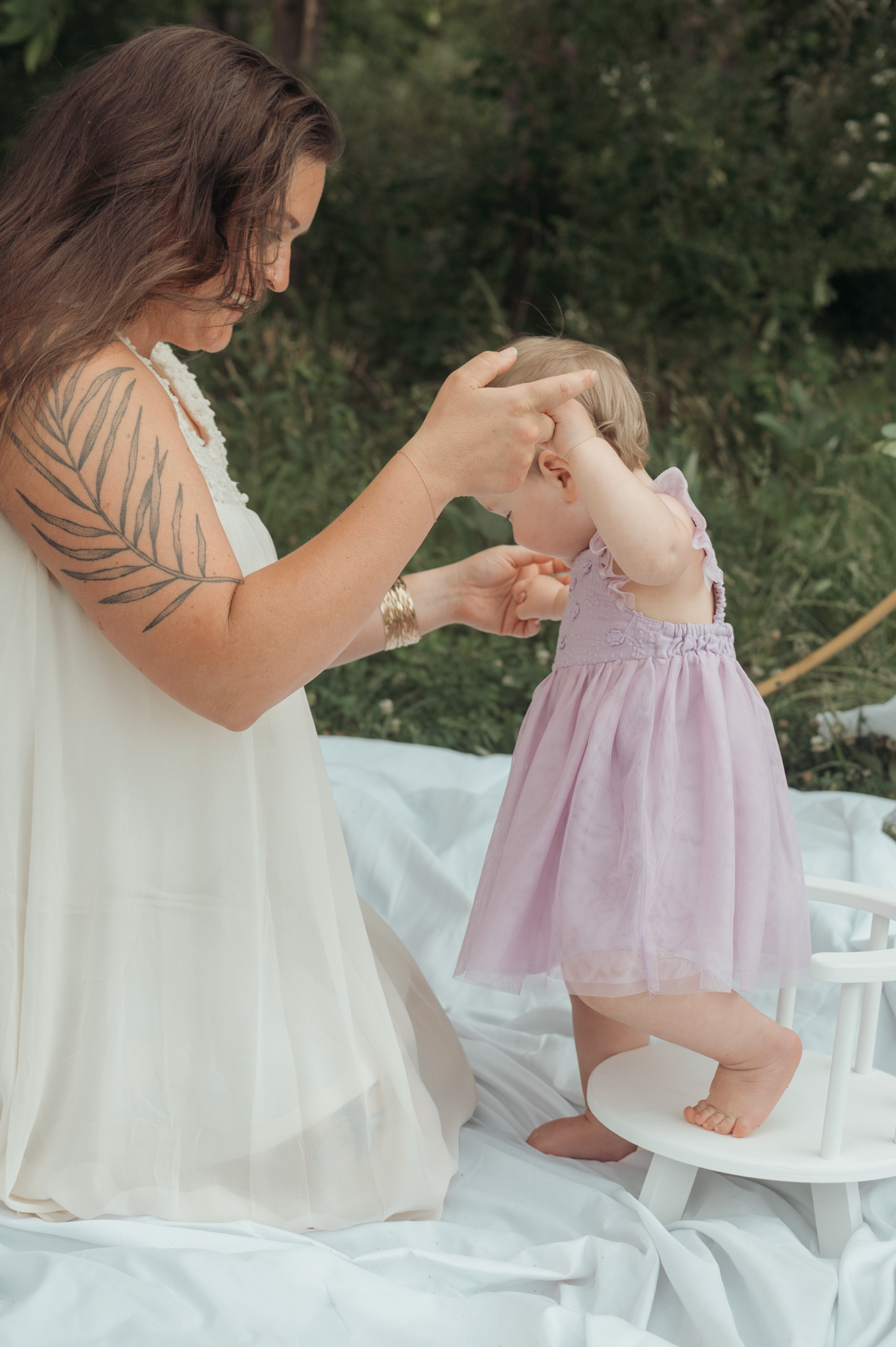 A tender moment as someone helps a small child in a pink dress balance on a white blanket outdoors.