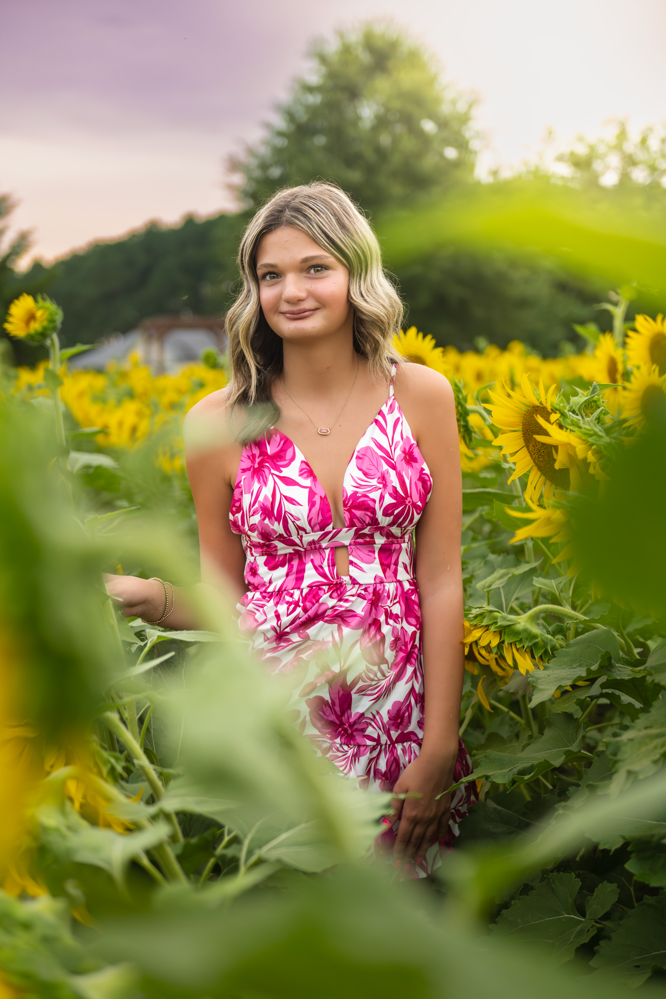 Pink floral sundress in a bright sunflower field during golden hour with blurred green foliage in the foreground.