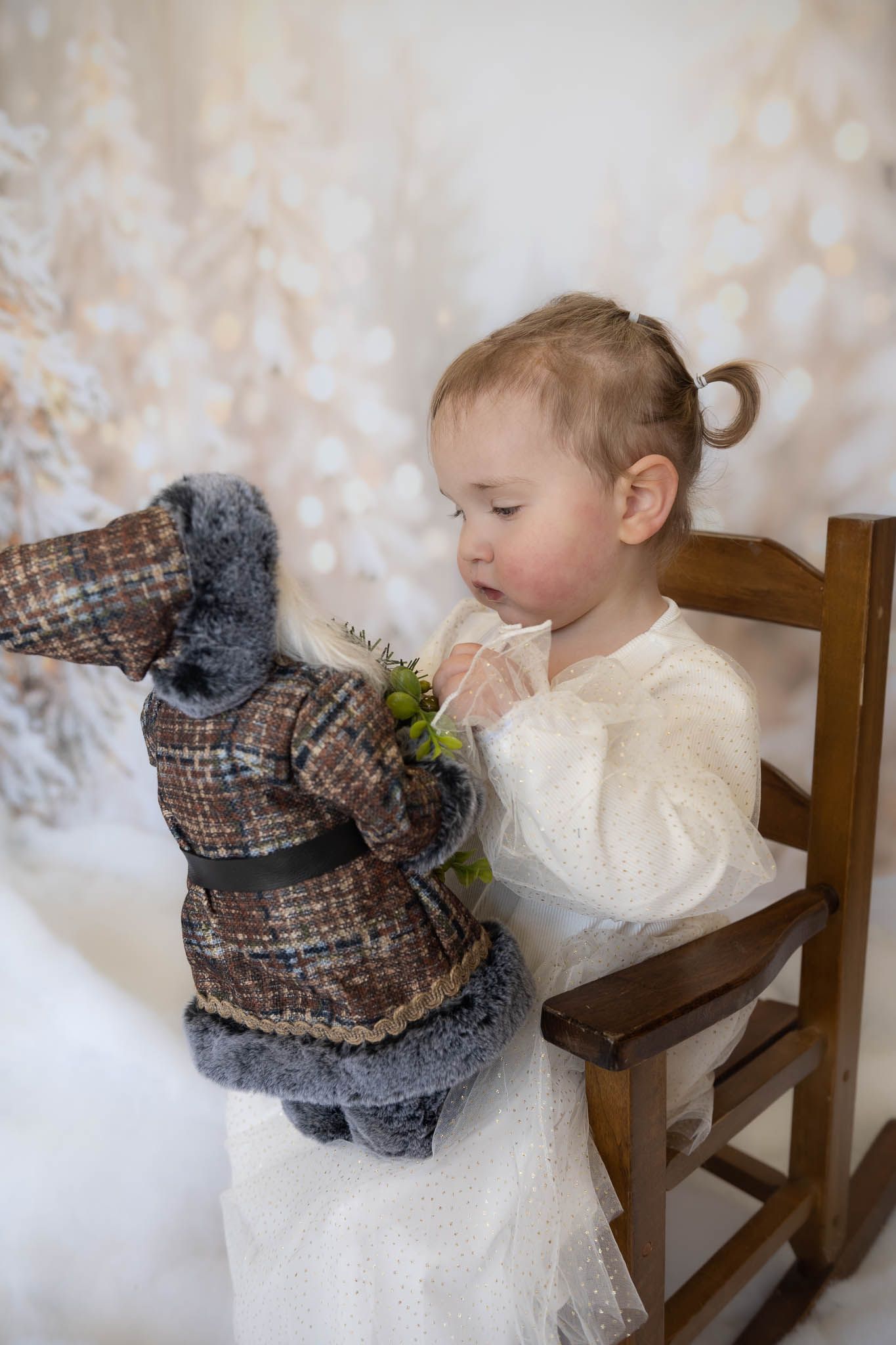 A small child sits in a wooden chair interacting with a plush owl toy against a soft white background.
