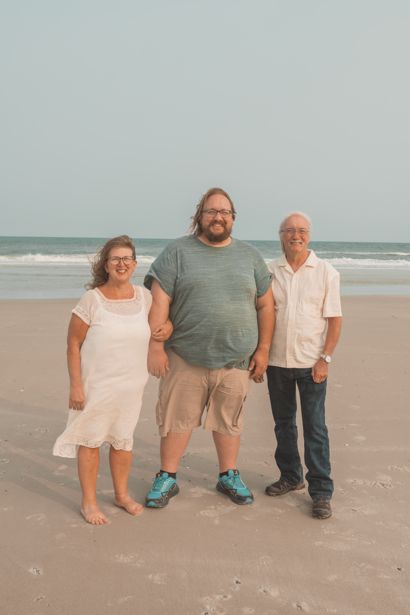Three people standing together and smiling on a sandy beach with ocean waves in the background.