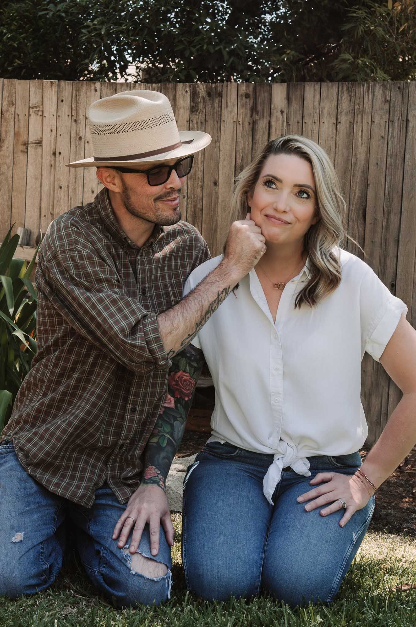 A couple sits together outdoors against a wooden fence, sharing playful moments in casual attire.