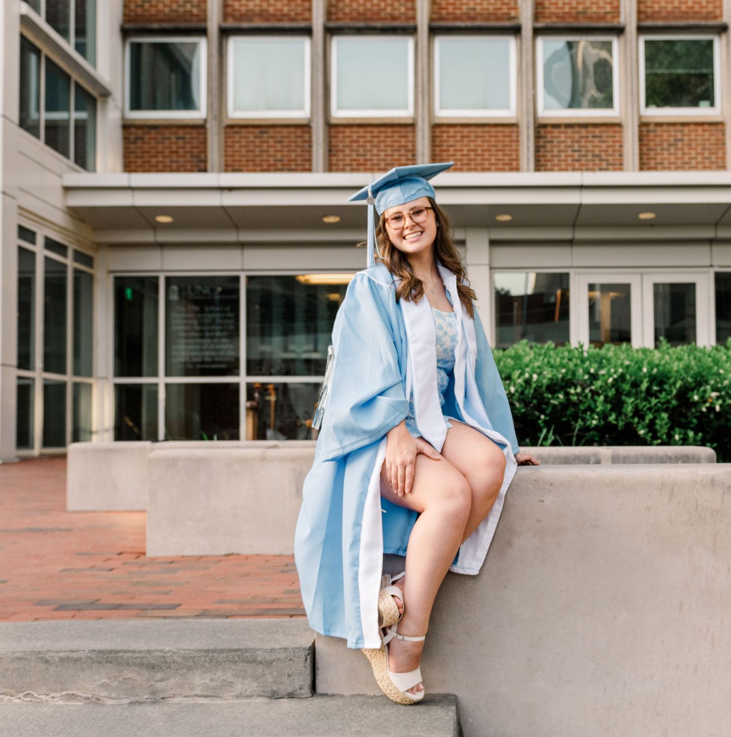 A graduate in light blue regalia and graduation cap sitting casually on a brick building ledge during commencement.