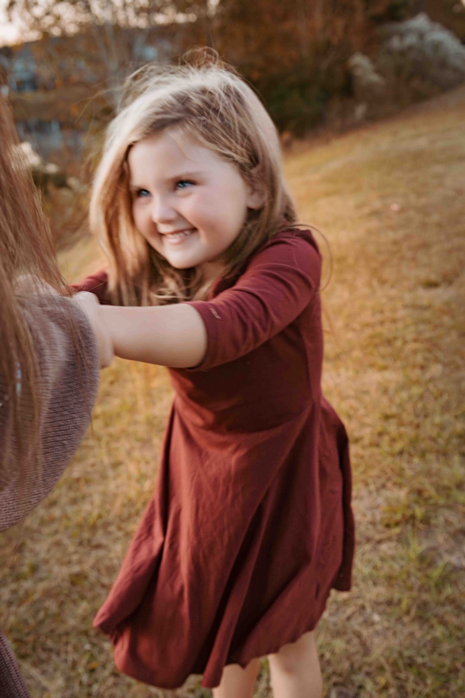 A child in a burgundy dress smiles while playing outdoors on an autumn day.