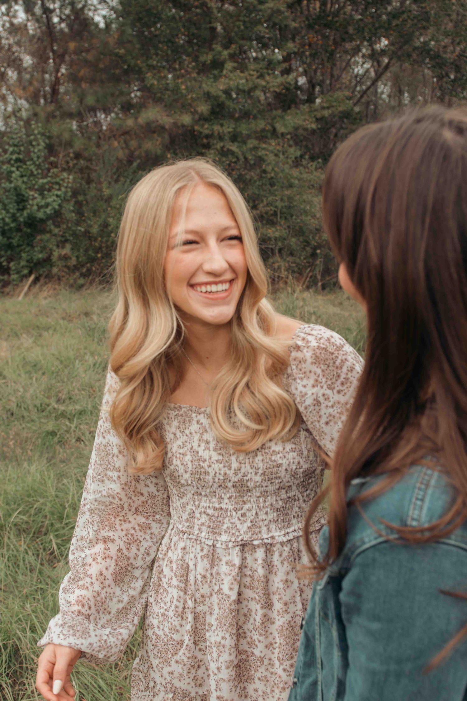 Two friends share a joyful moment outdoors, one wearing a floral dress and the other in denim against a natural backdrop.