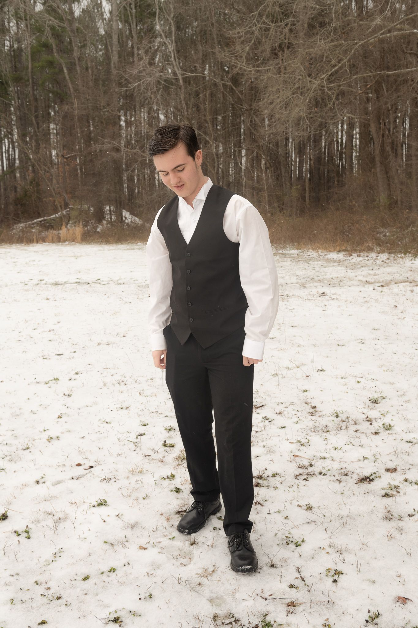 Person in formal attire with black vest and white dress shirt stands thoughtfully in snowy winter landscape.