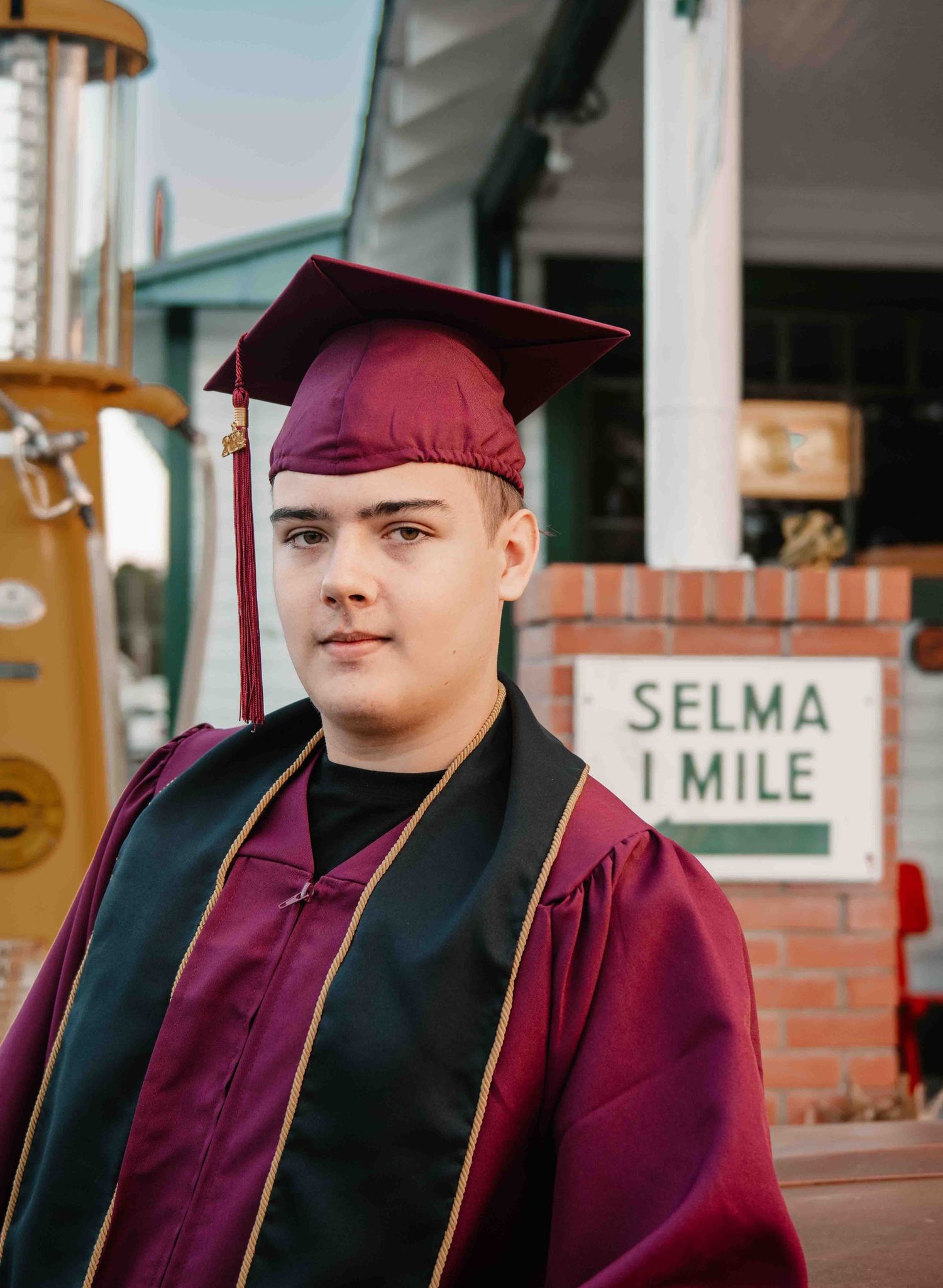 A graduate in burgundy cap and gown stands beside a Selma 1 Mile sign.