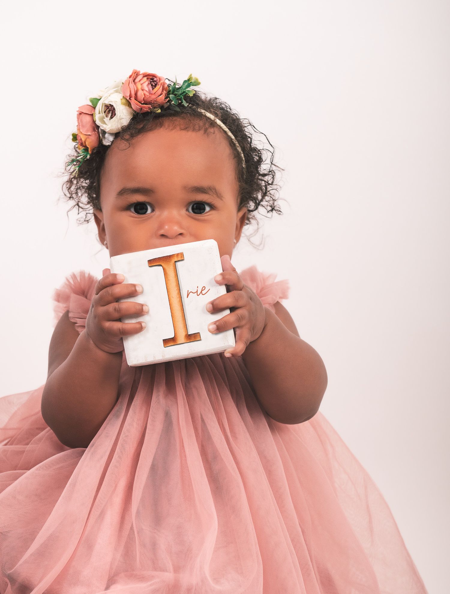 A child in a pink dress holds alphabet blocks and wears a floral crown for an artistic milestone photo session.