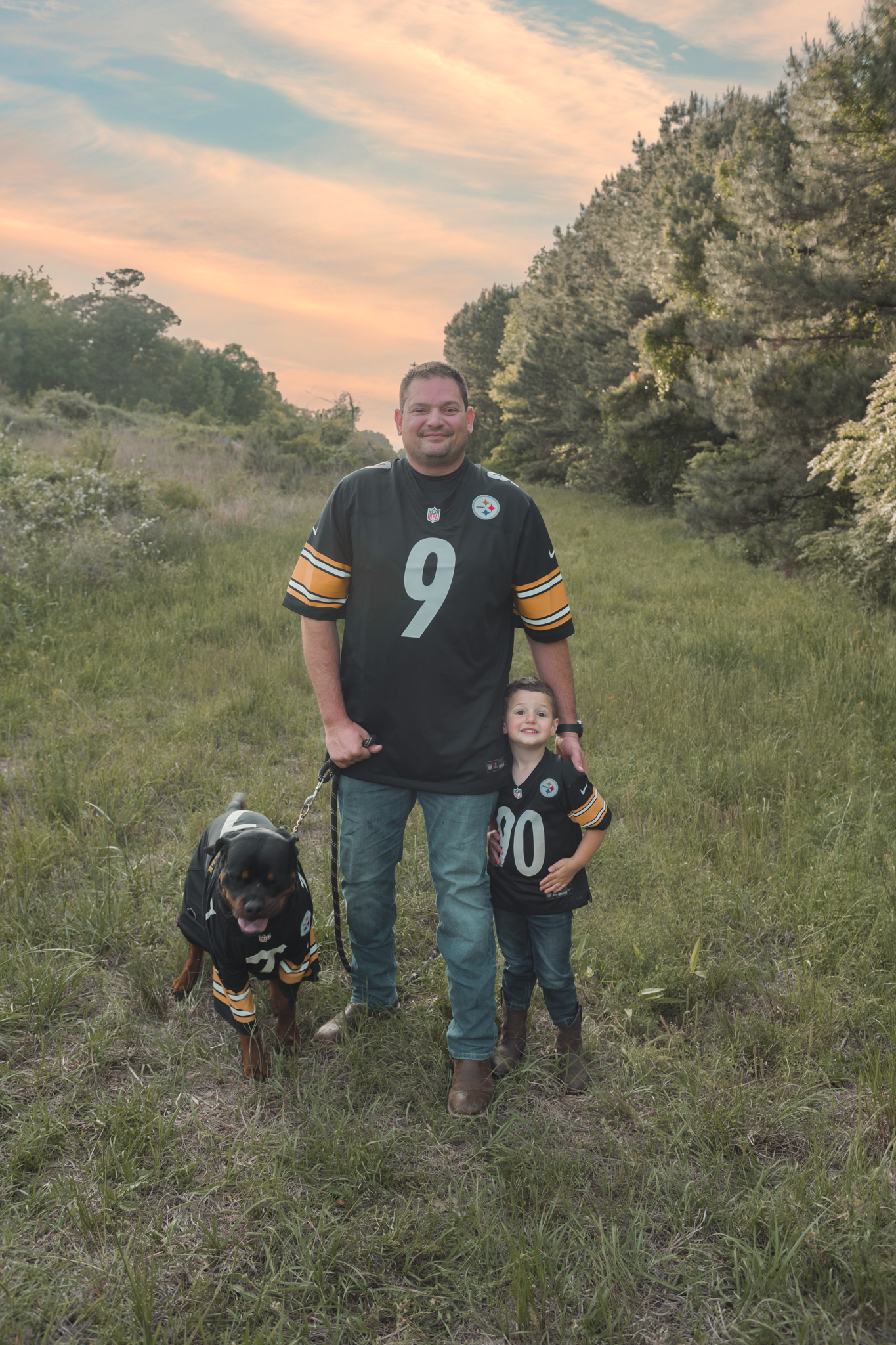 Pittsburgh Steelers fans stand in a grassy field with their dog at sunset against a backdrop of pine trees.