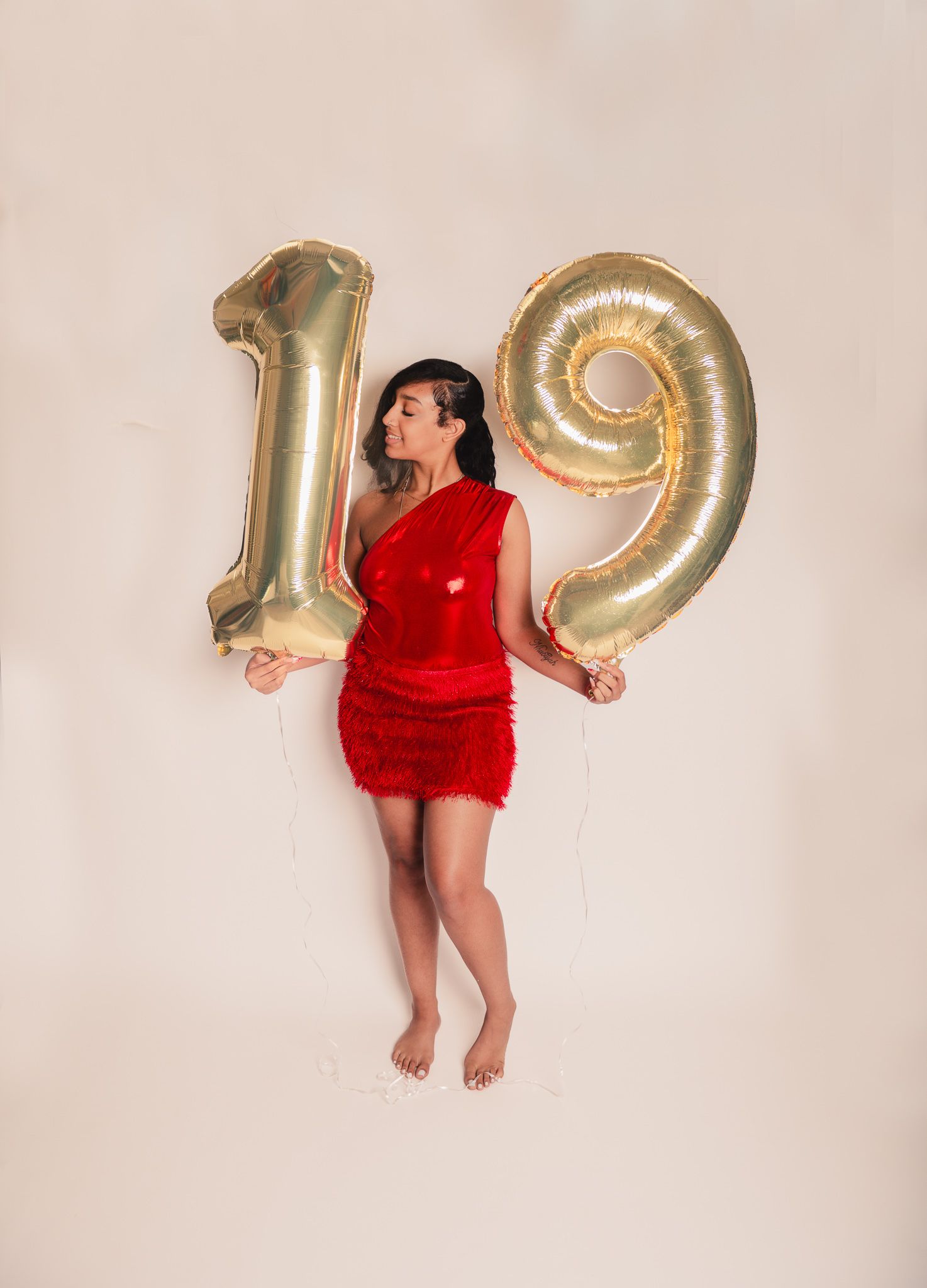 A person in a red mini dress poses with large gold number balloons showing 19 against a light pink background.