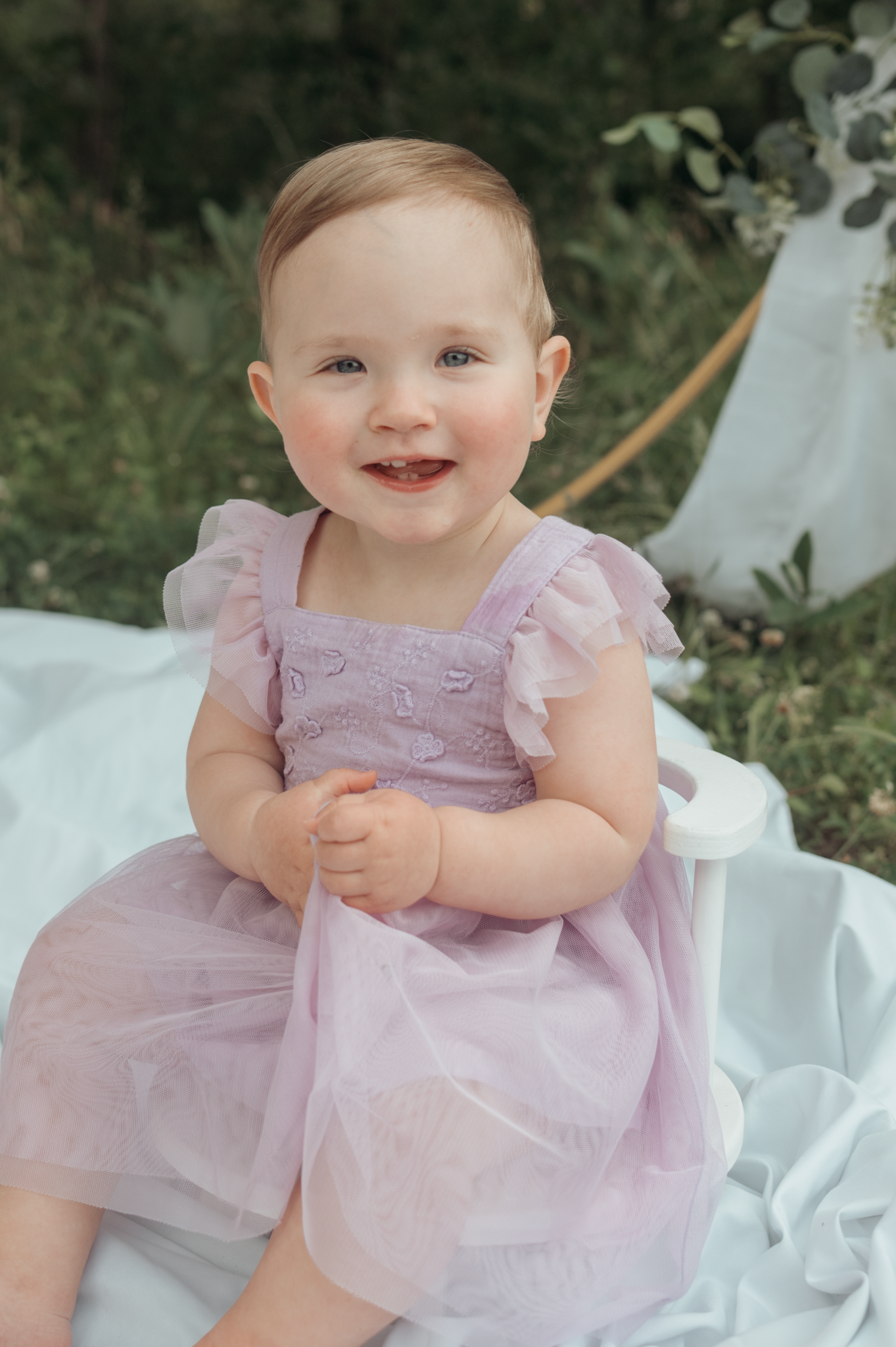 A smiling baby wearing a lavender tulle dress sits on a white blanket in an outdoor garden setting.