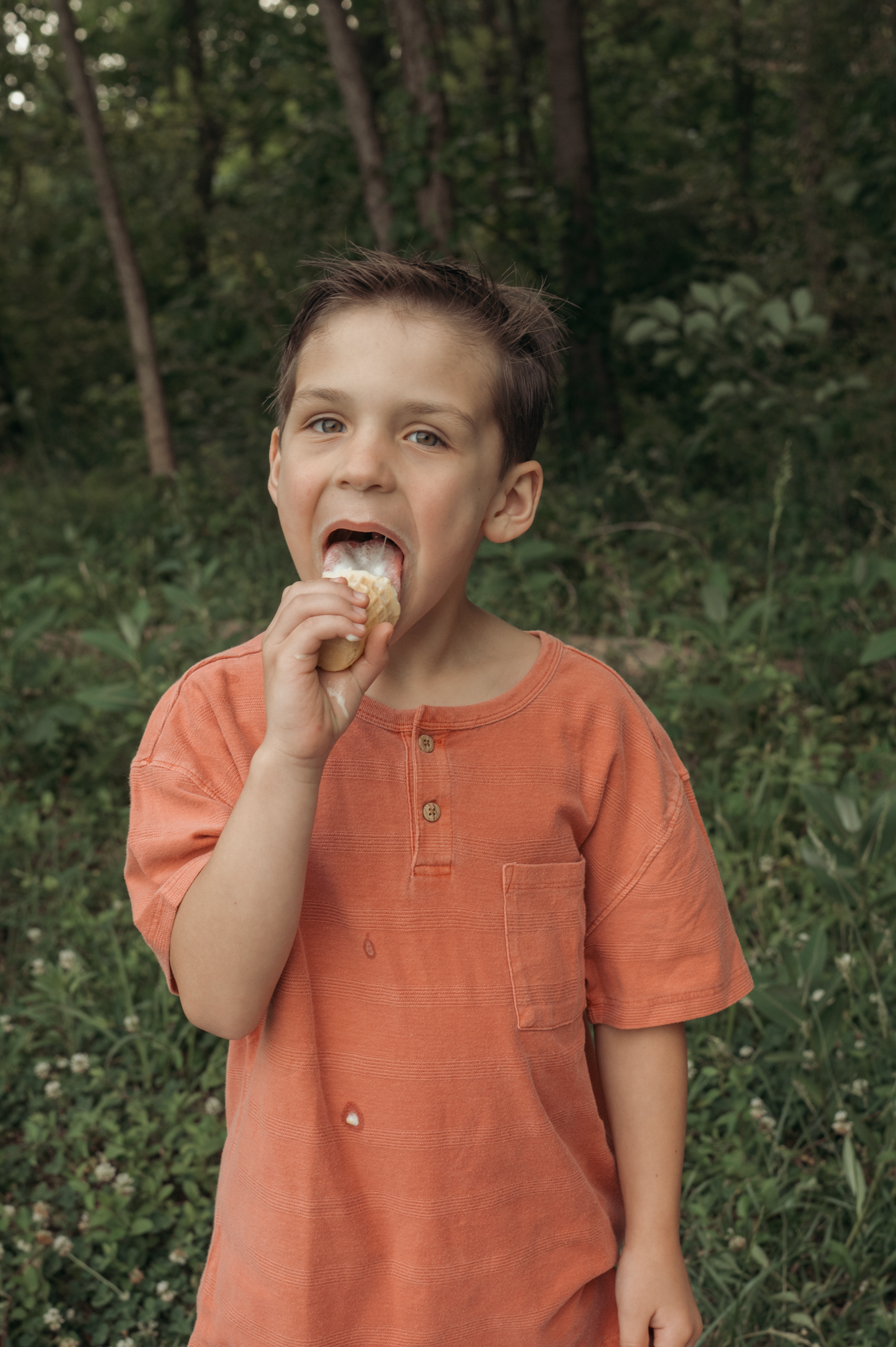 A child in an orange t-shirt takes a bite of food while standing in a wooded outdoor setting.