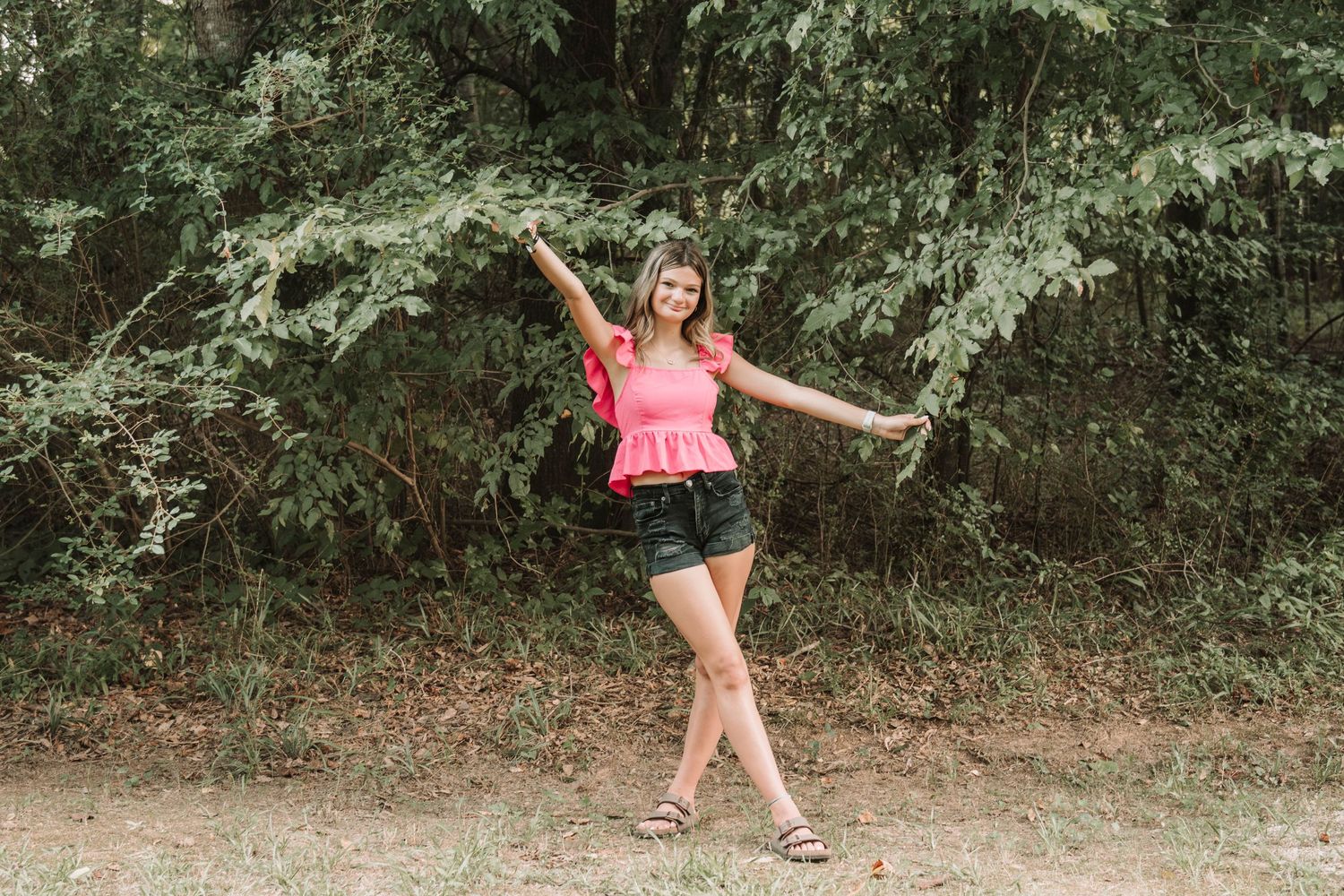 Someone in a pink tank top and denim shorts strikes a playful pose on a wooded dirt path on a summer day.