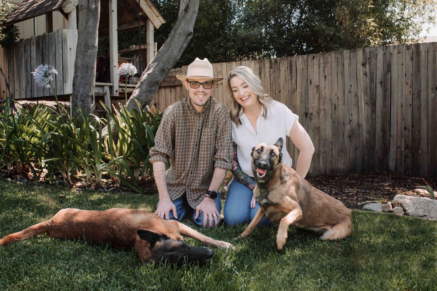 Couple in casual western attire sitting on grass with two dogs in backyard setting with wooden fence.
