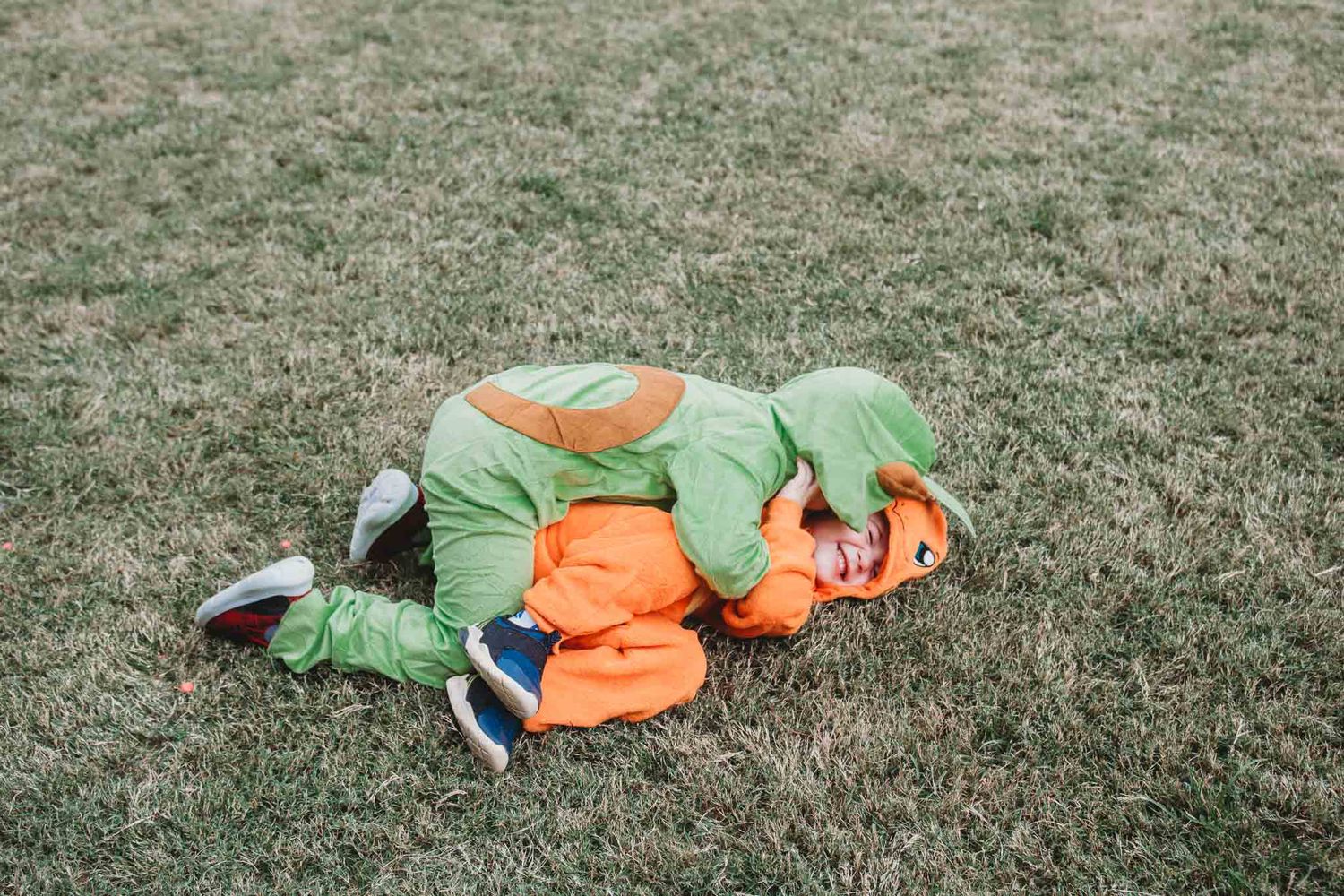 A person dressed in a green and orange mascot costume lies face down on grass.