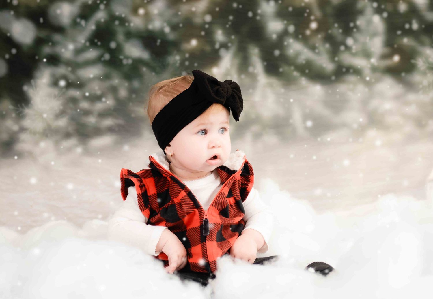 A baby wearing a buffalo plaid vest and black headband poses in a winter wonderland setting with falling snow.