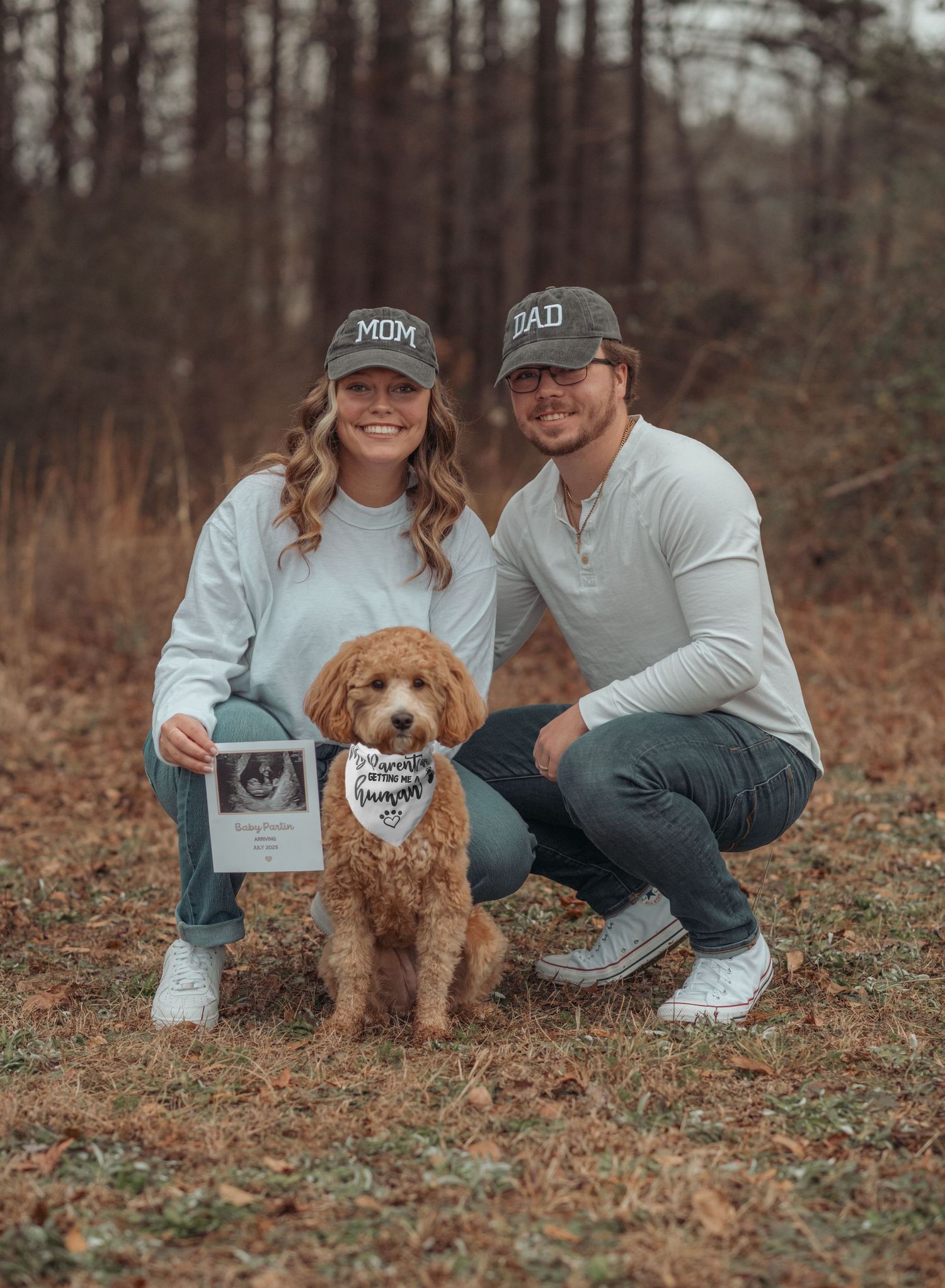 Through autumn woods, an adorable goldendoodle dog poses with announcement sign and ultrasound photo with mom & dad in Smithfield, NC