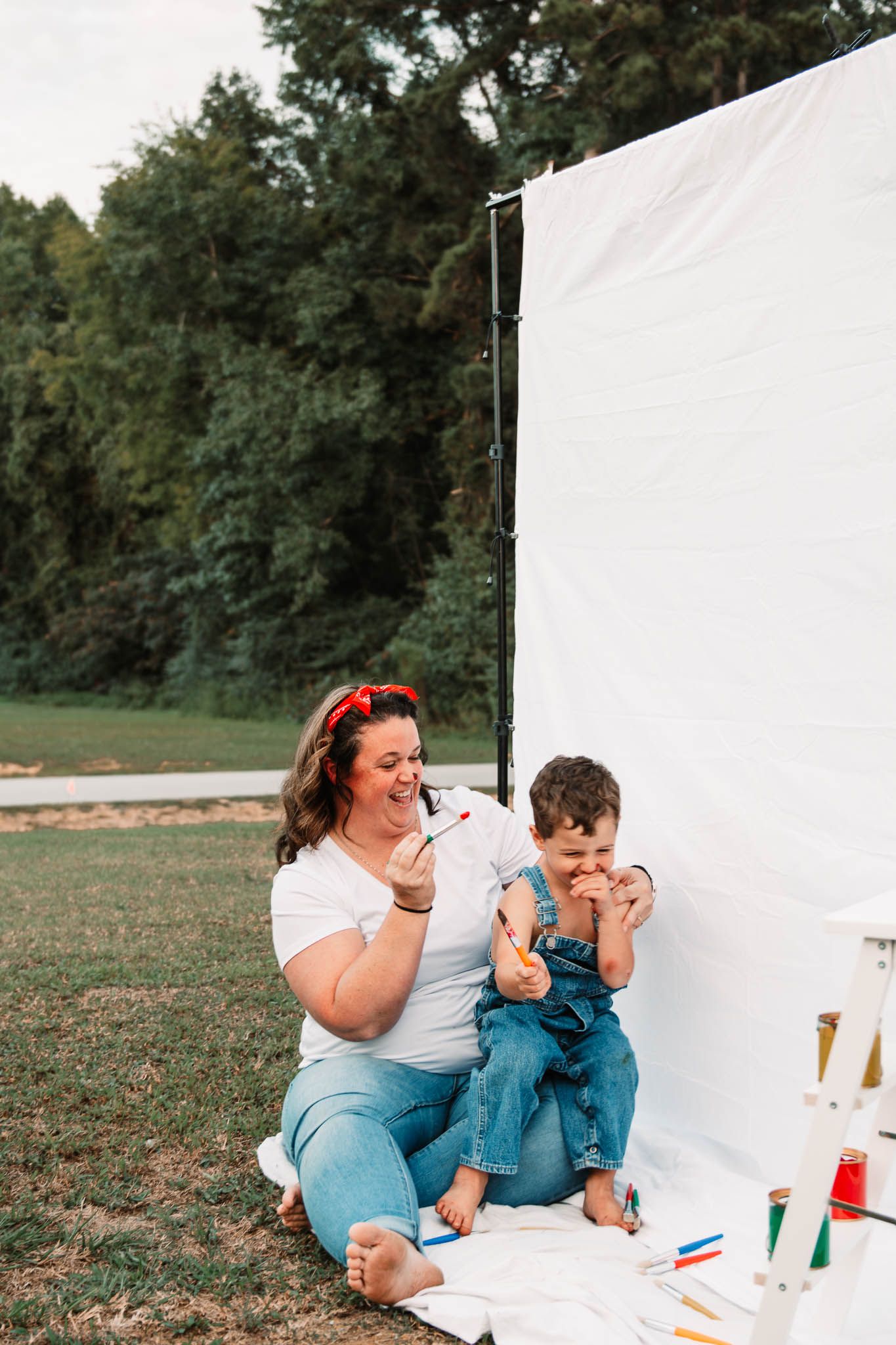 Parent and child enjoy painting time together on a white sheet outside surrounded by art supplies.