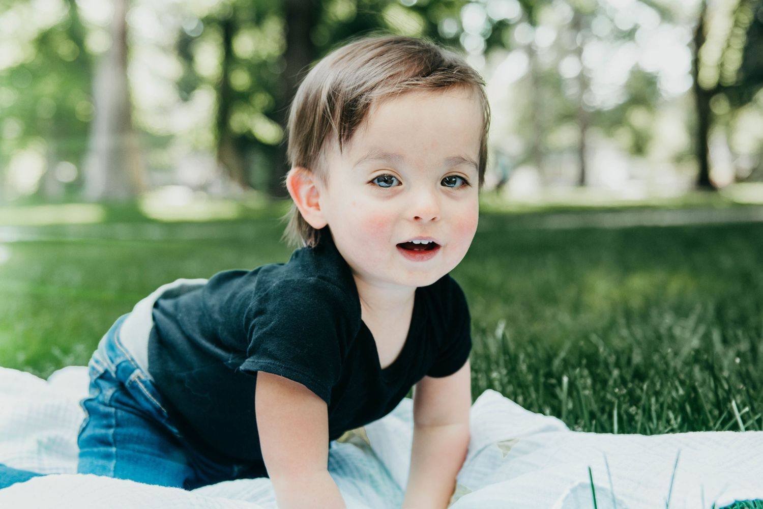 A small child wearing a black t-shirt and jeans crawls on a white blanket in the grass on a sunny day.