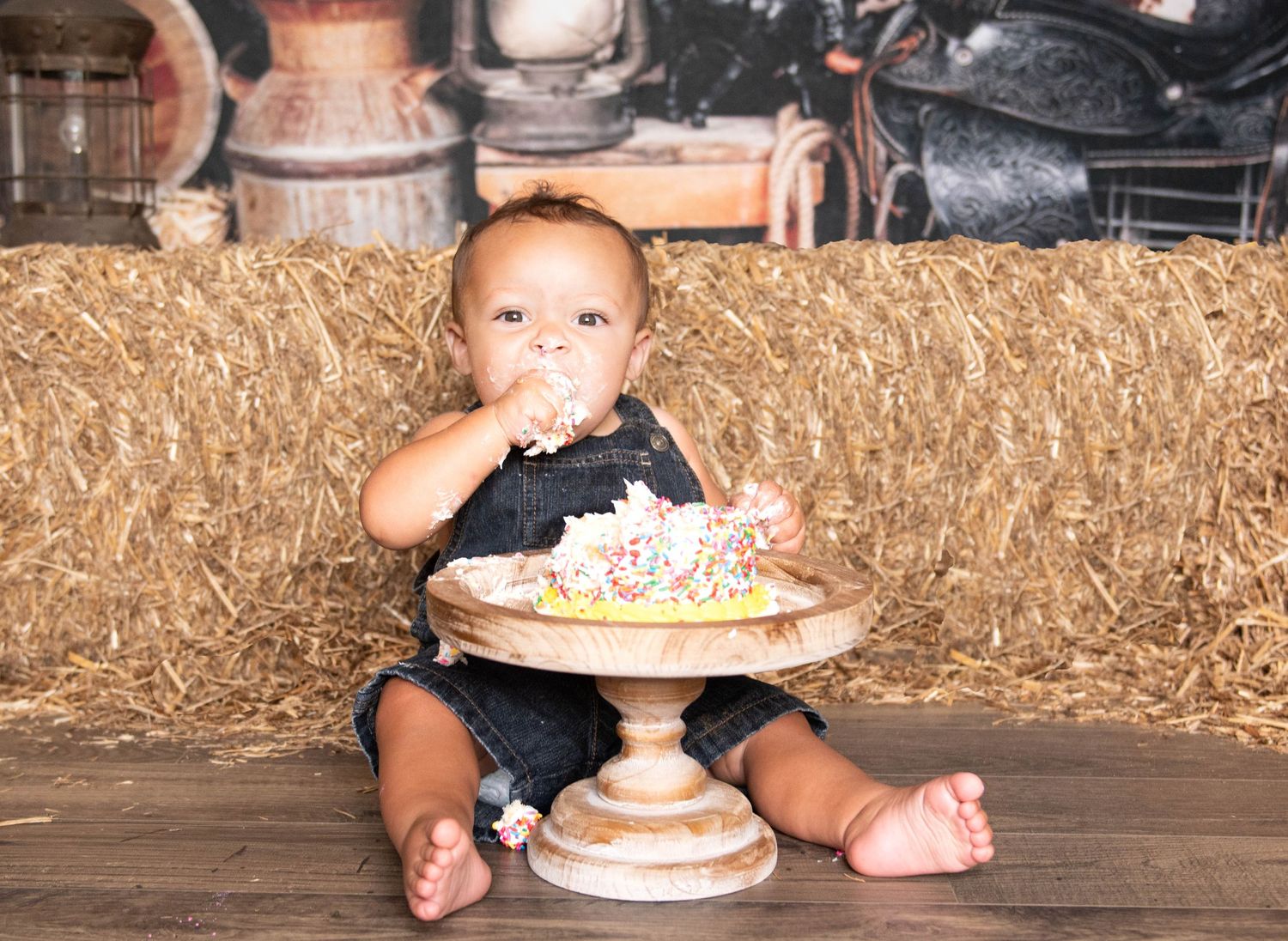 Small child in overalls sitting on wooden pedestal with cake enjoying messy birthday cake smash photo session.