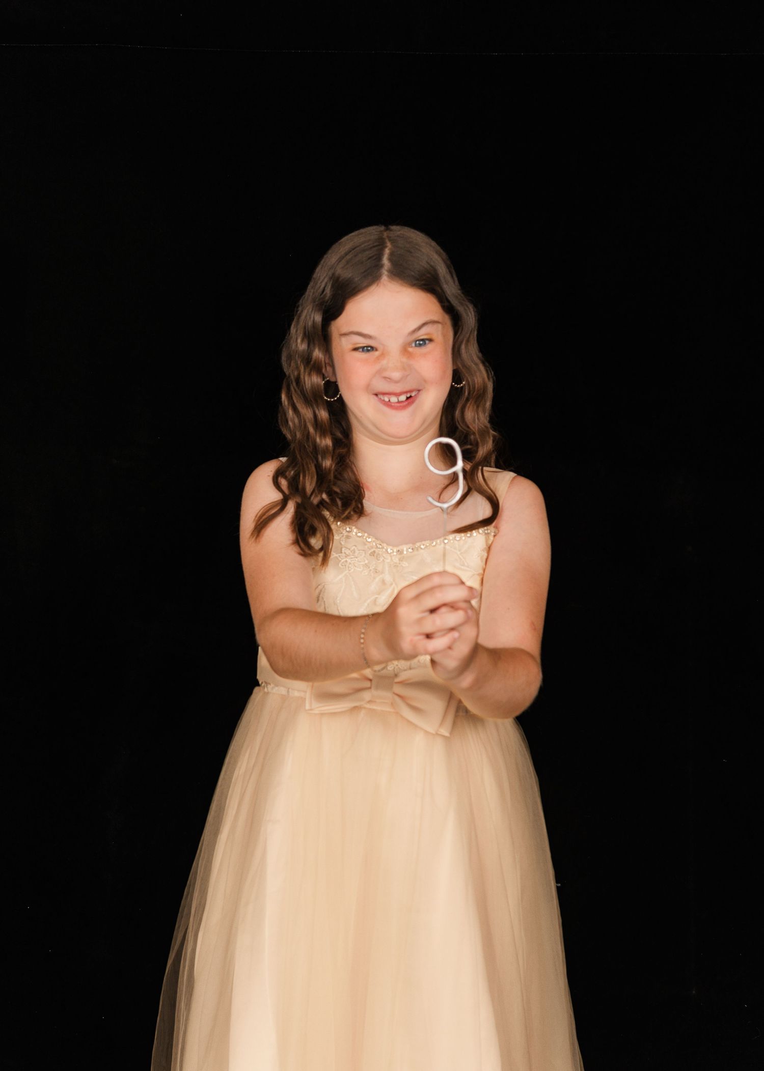 A young person in a cream-colored formal dress smiles and laughs while holding something against a dark background.