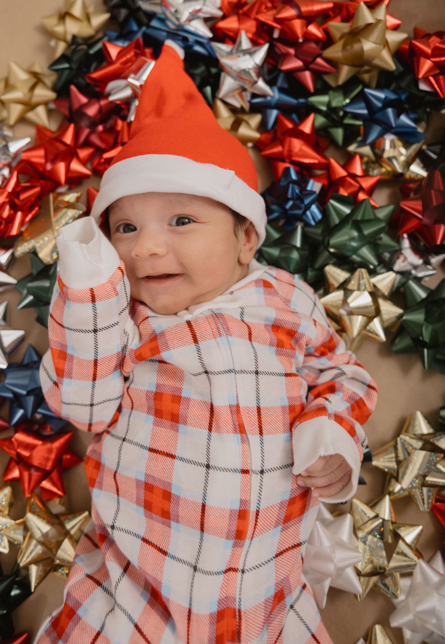 A baby wearing a red and white plaid outfit and Santa hat lies on a festive background of colorful gift bows.