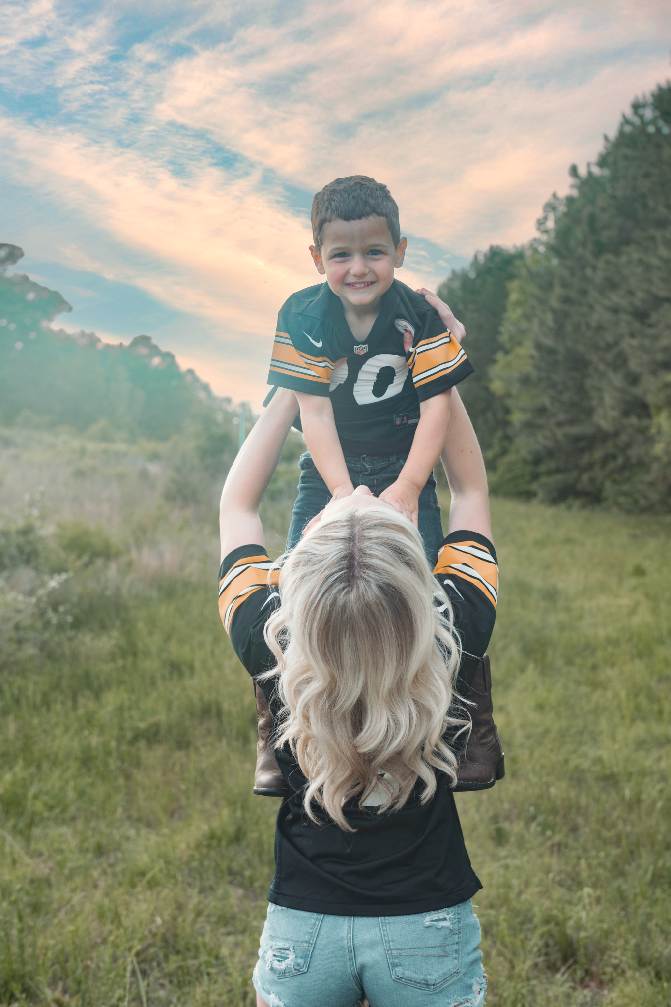 A young football fan in a green jersey is lifted up playfully in a grassy field during sunset.