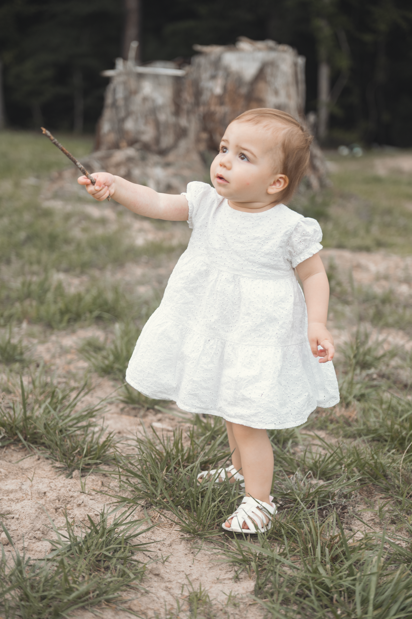 A small toddler in a white lace dress holds a stick while standing outside in grass near a rustic structure.