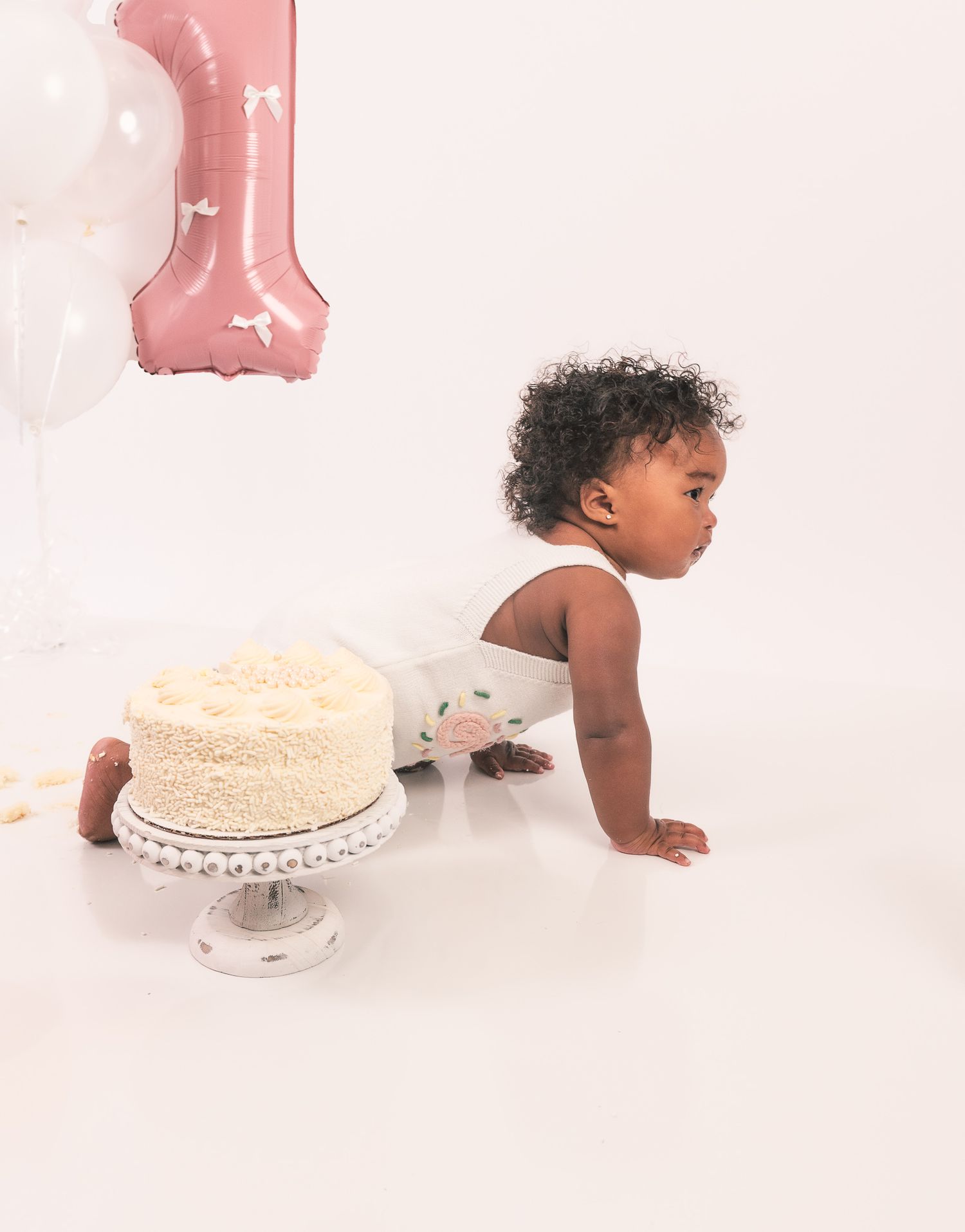 Baby in diaper celebrates first birthday with white cake and pink balloon decorations on minimalist set.