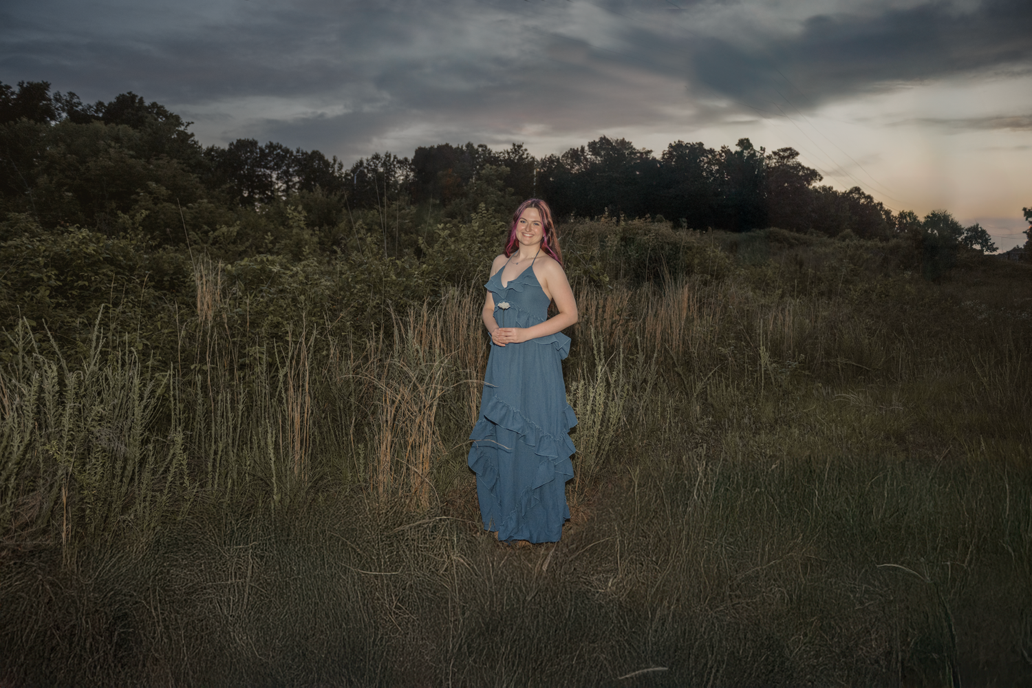 Haylee in a blue dress stands amid tall grass during sunset with dramatic cloudy skies in the background during her senior session