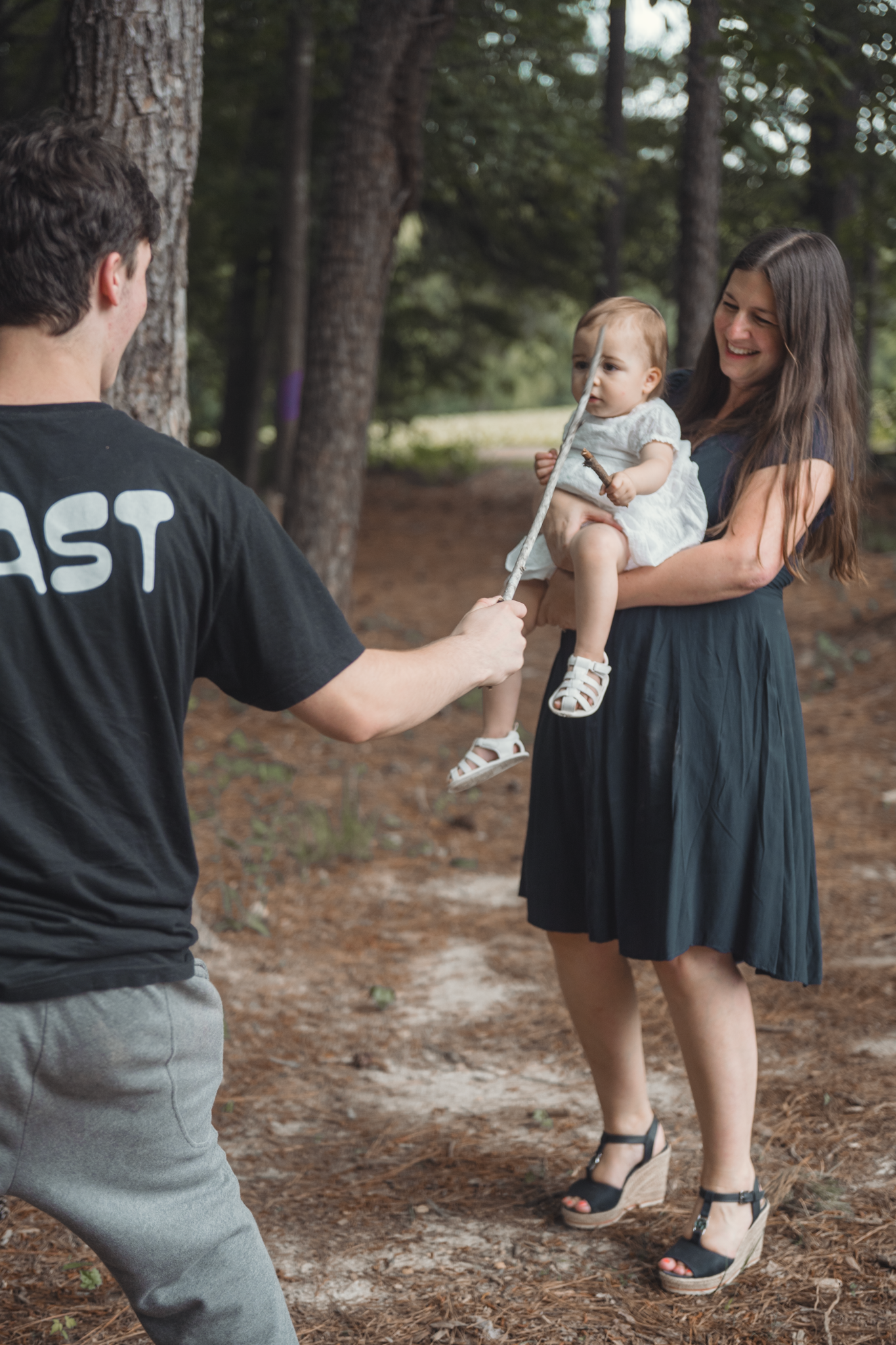 A parent in a dark t-shirt interacts with their small child being held in a forested outdoor setting.