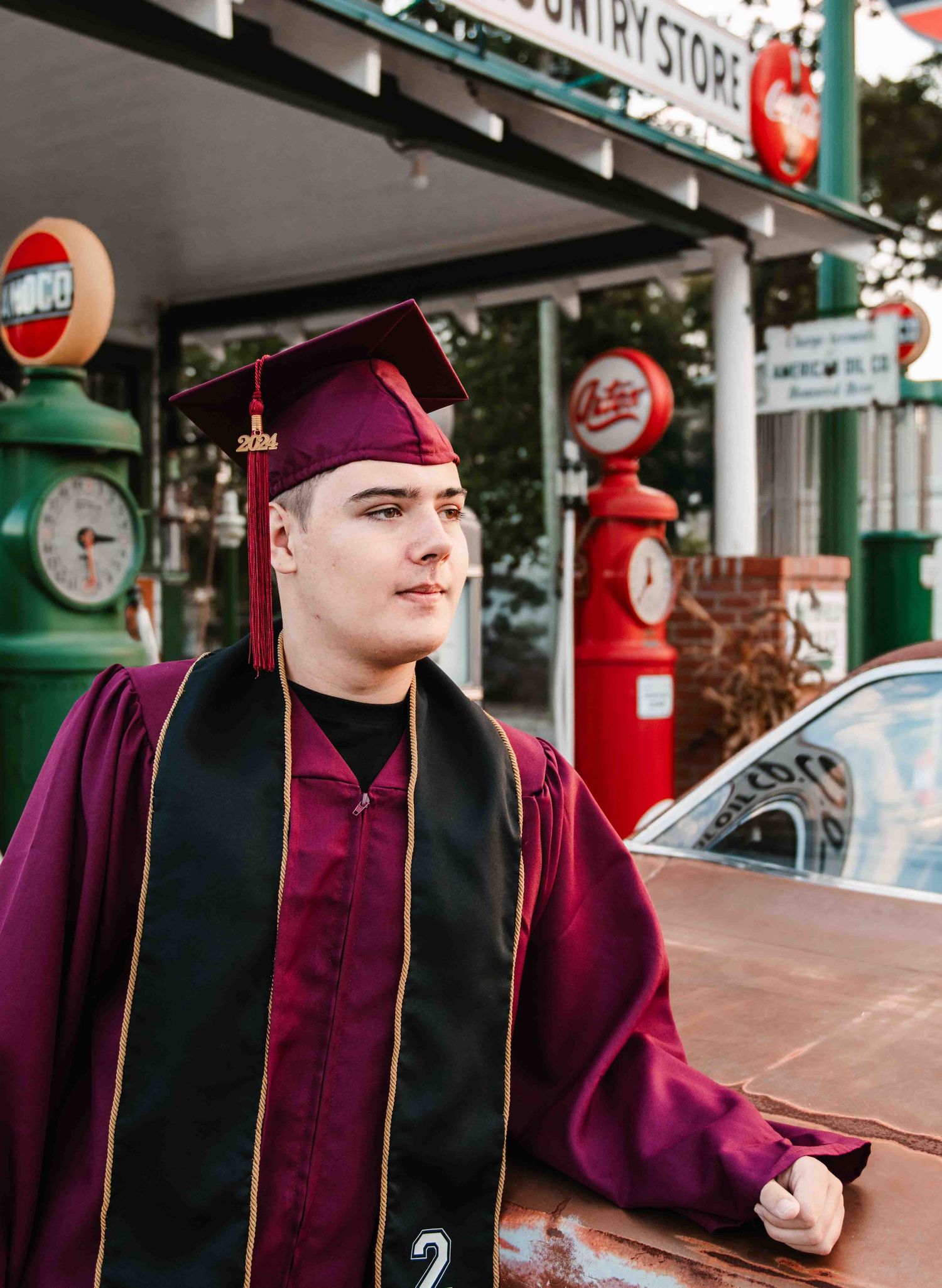 High school graduate in burgundy cap and gown poses at retro gas station with vintage signs in Smithfield, NC