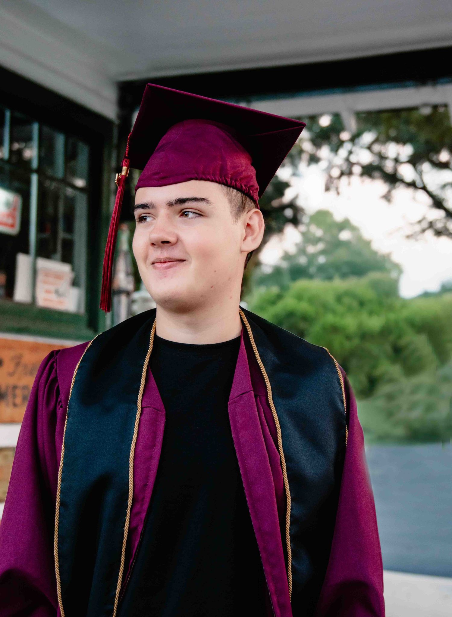 Graduate in maroon and black cap and gown stands outdoors looking to the side with a slight smile.
