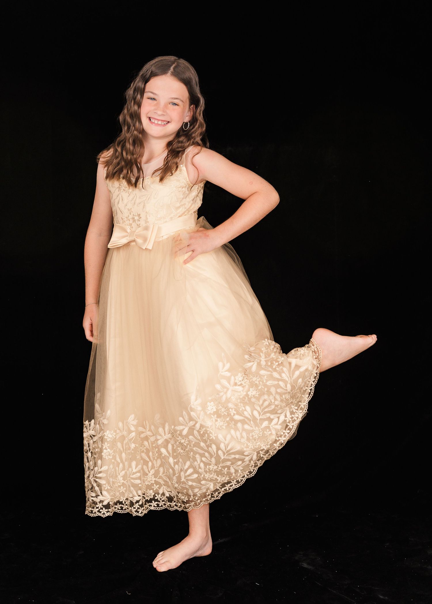 A model poses in an elegant champagne-colored formal dress with lace details against a black background.