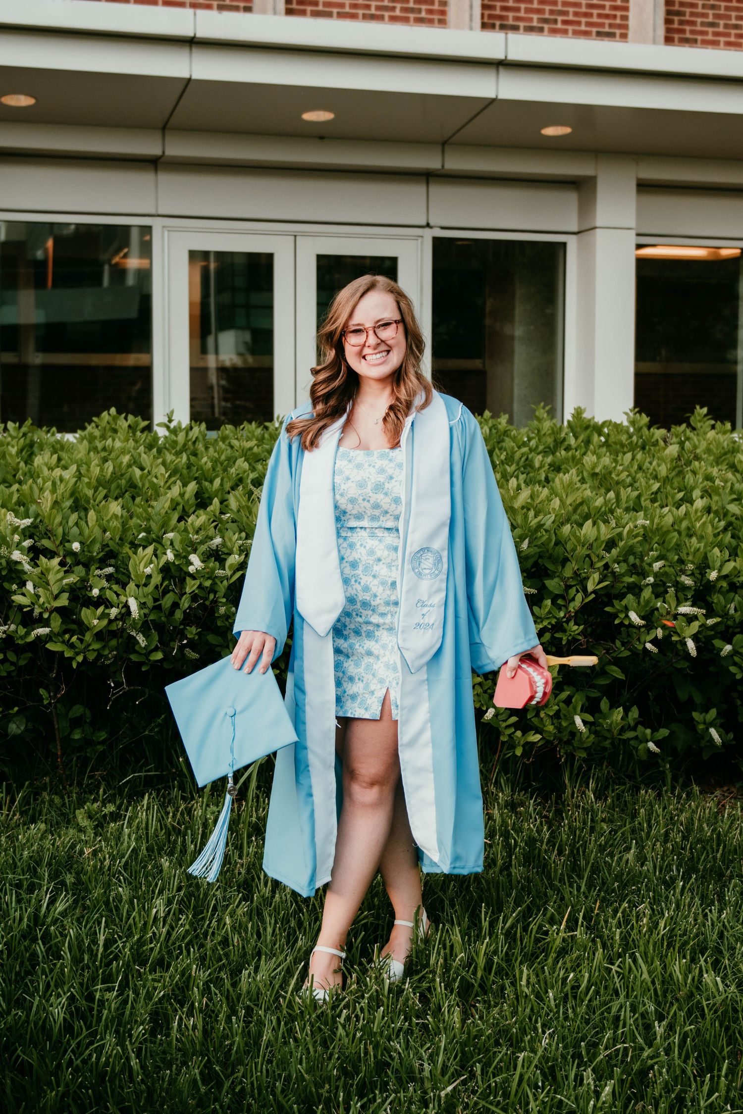 A graduate stands on grass wearing a light blue graduation gown and cap while holding a diploma in front of a brick building.